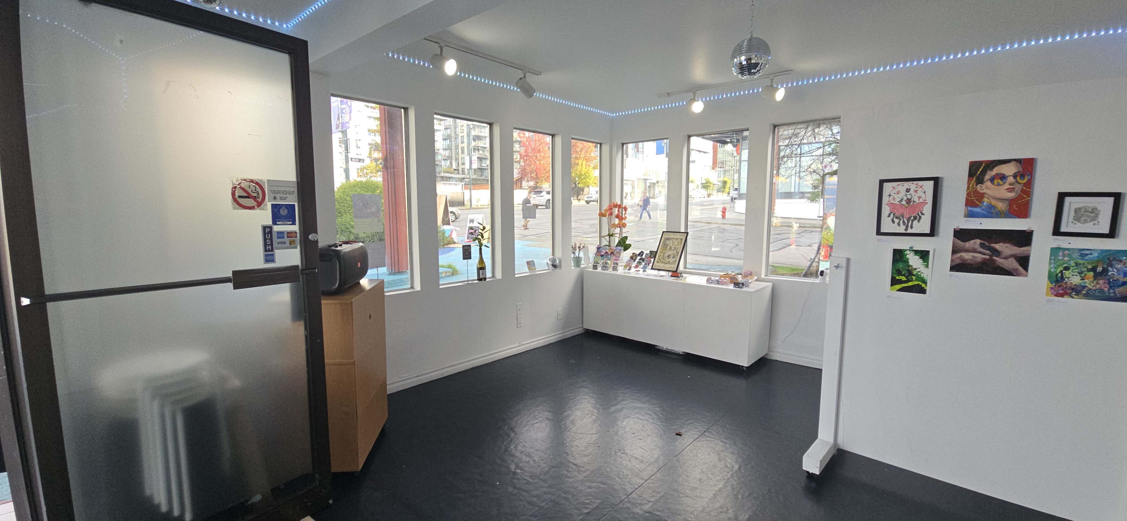 The image shows a bright, well-lit interior space featuring large windows, a display table adorned with various items, and art pieces hanging on the walls.