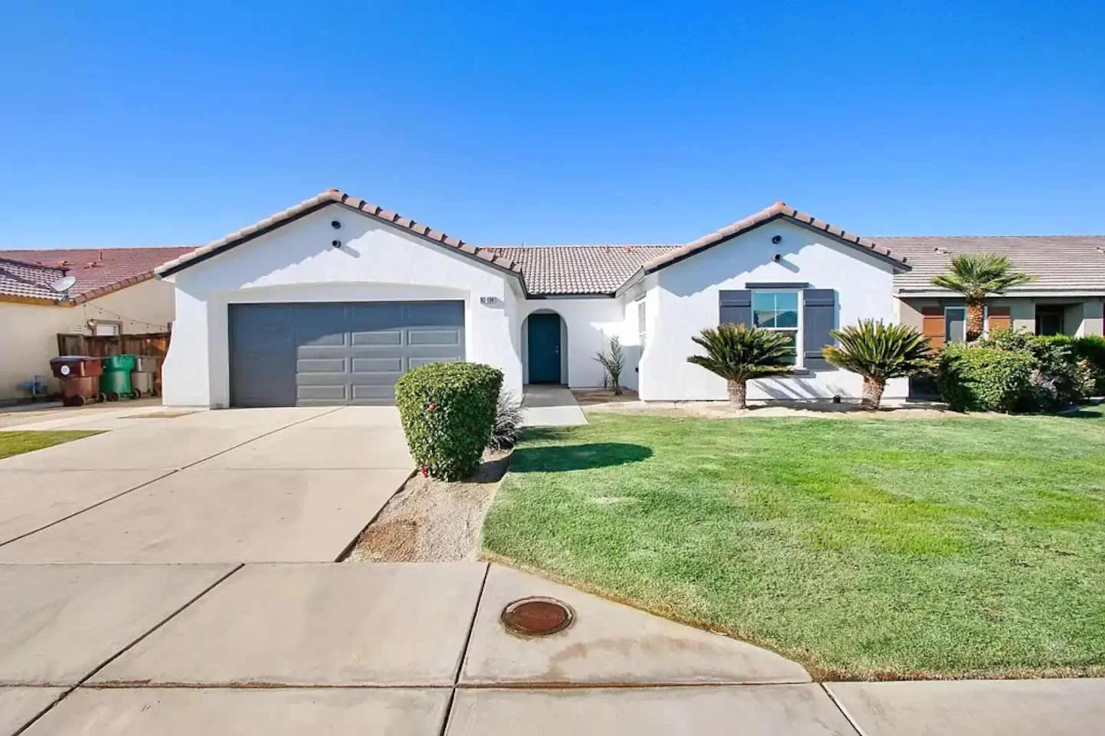 A single-story house with a gray garage door, green grass, and shrubs in the front yard under a clear blue sky.