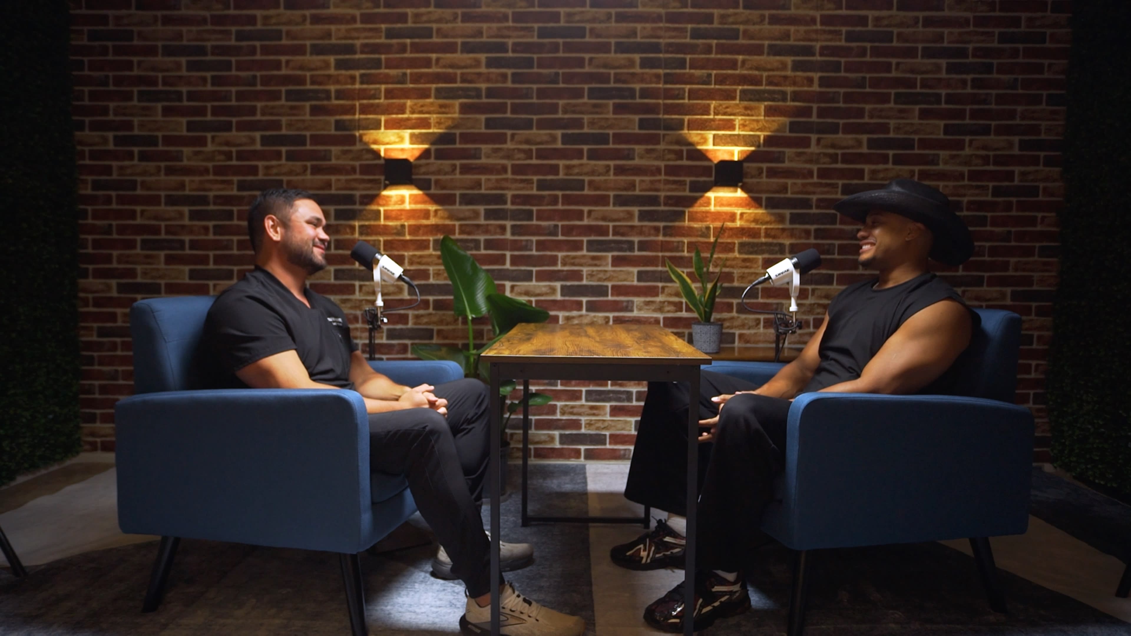 Two men sit in blue chairs across from each other at a wooden table, with brick walls and soft lighting in the background, equipped with microphones for a conversation.