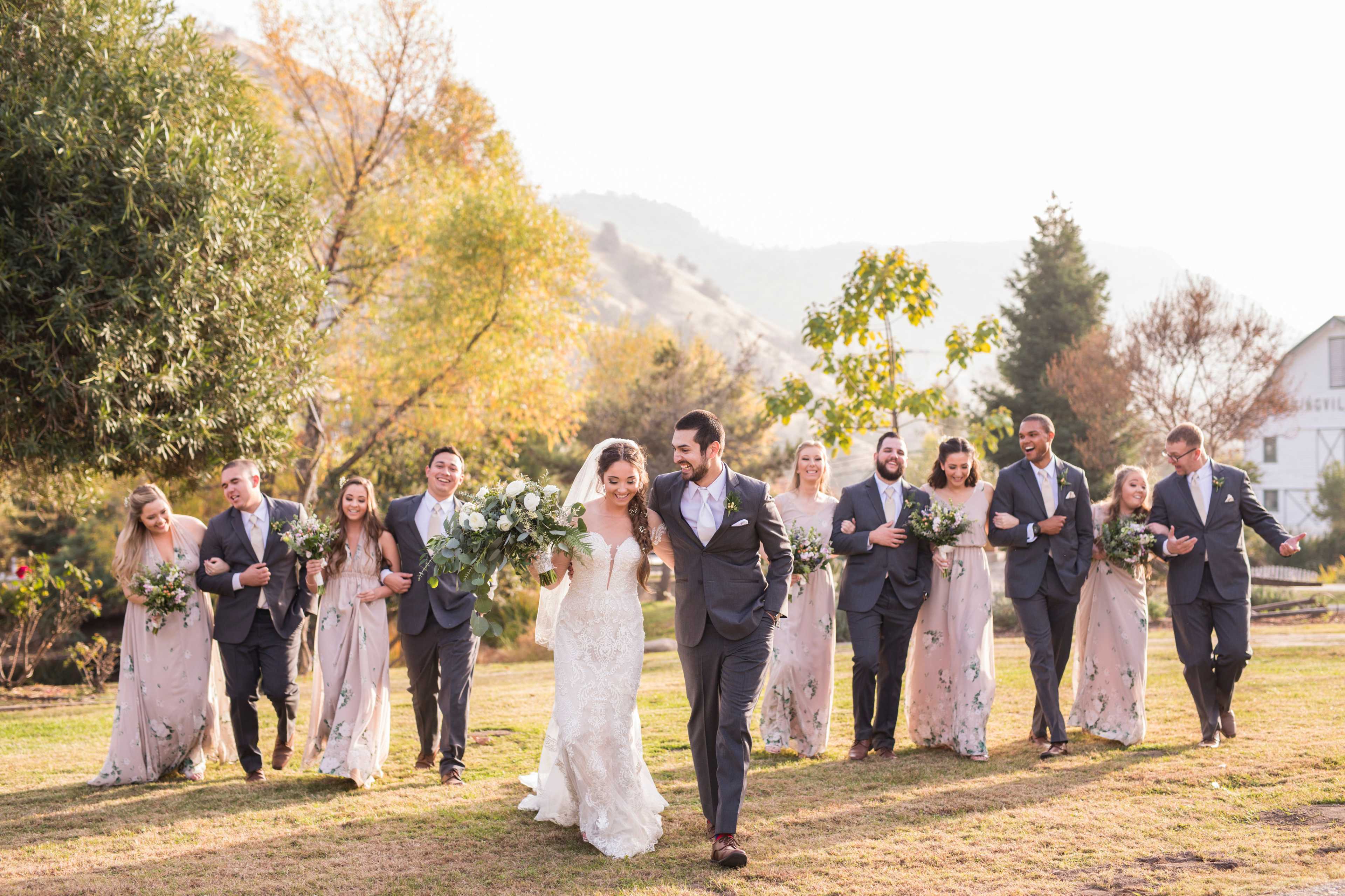 A wedding party walks together in a grassy outdoor setting, with the bride and groom leading the group.