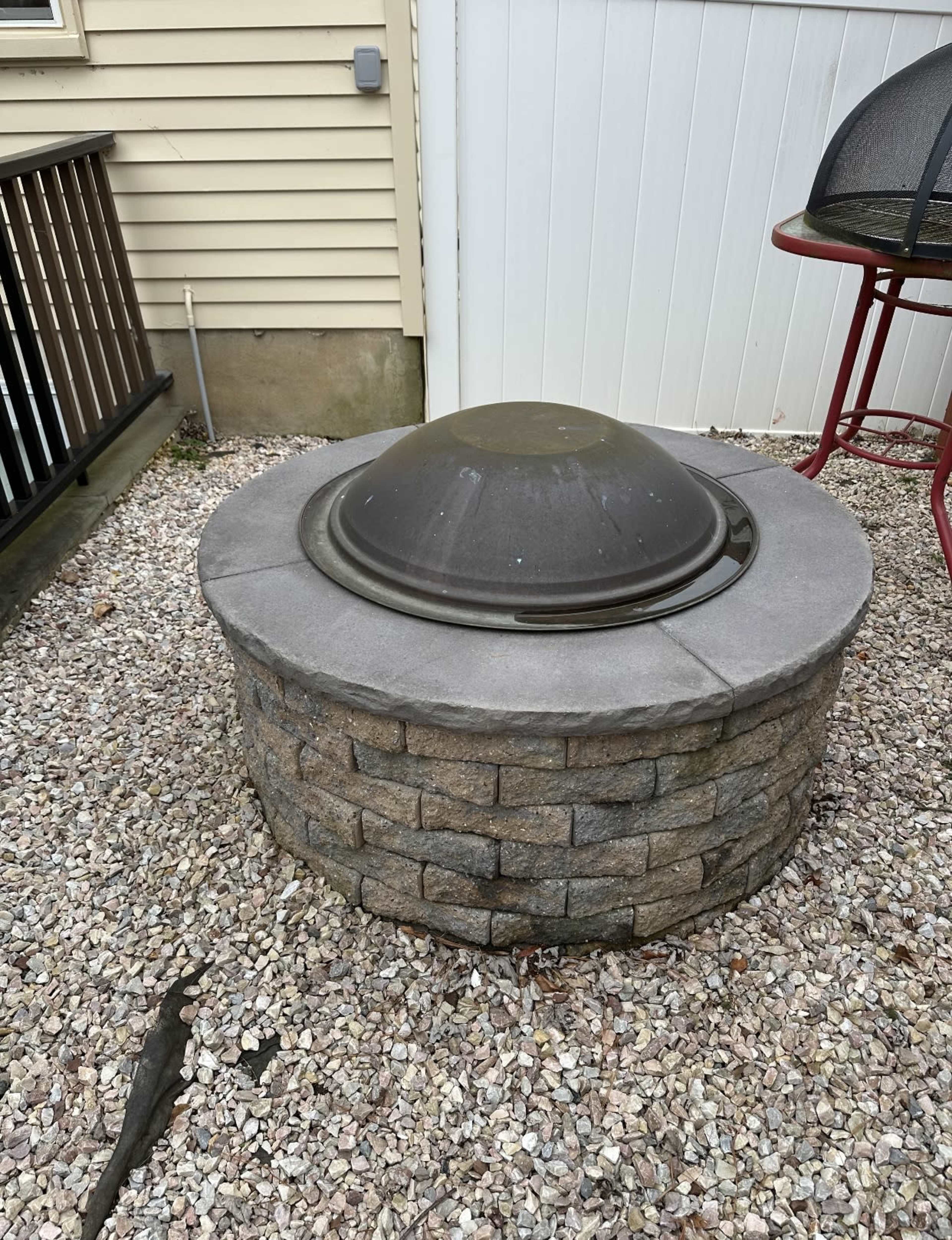 A stone fire pit with a metal lid situated on a gravel surface in a backyard.