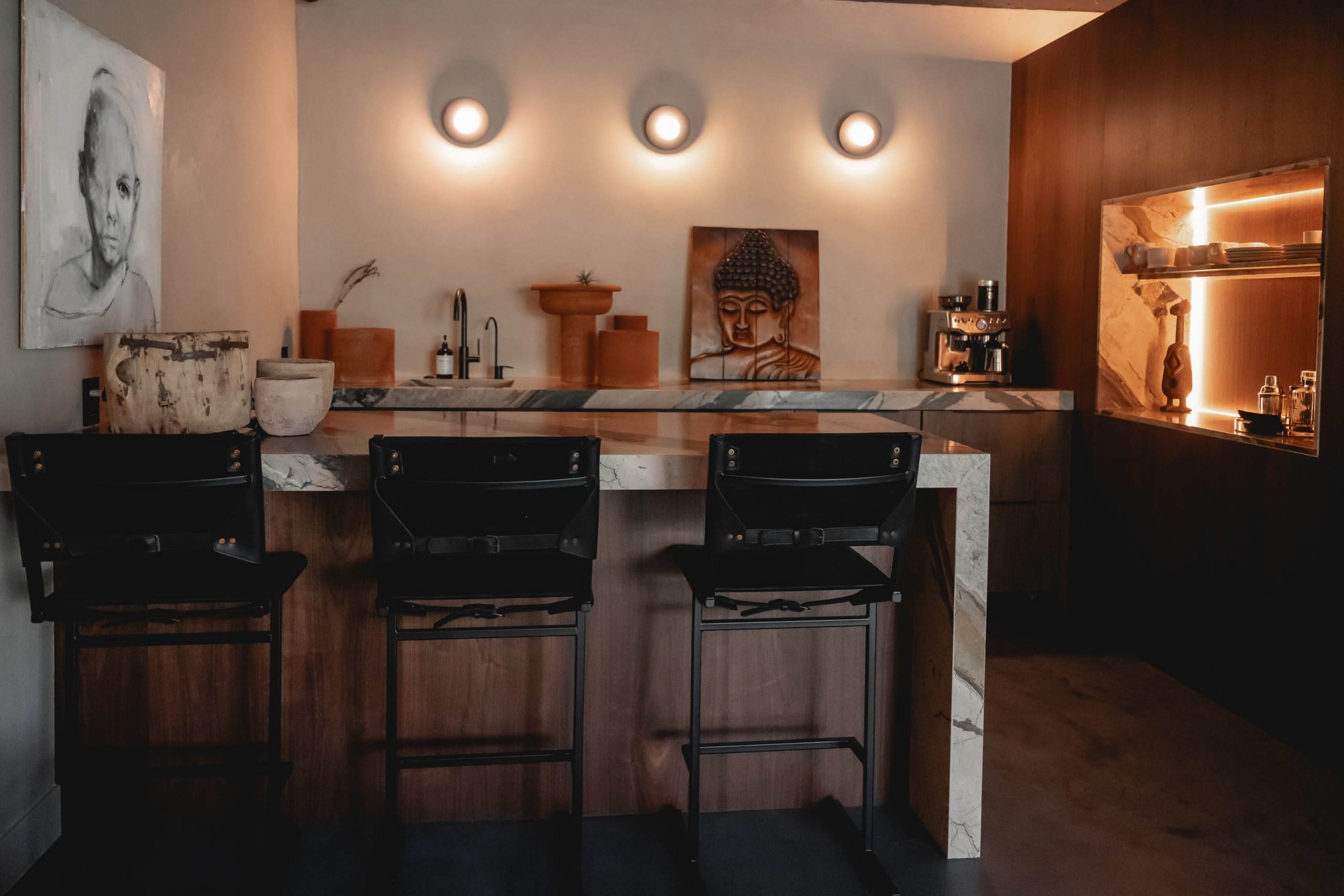 A modern interior of a kitchen bar area featuring three black chairs, a marble countertop, and a wall adorned with a Buddha sculpture and minimal decorative items.