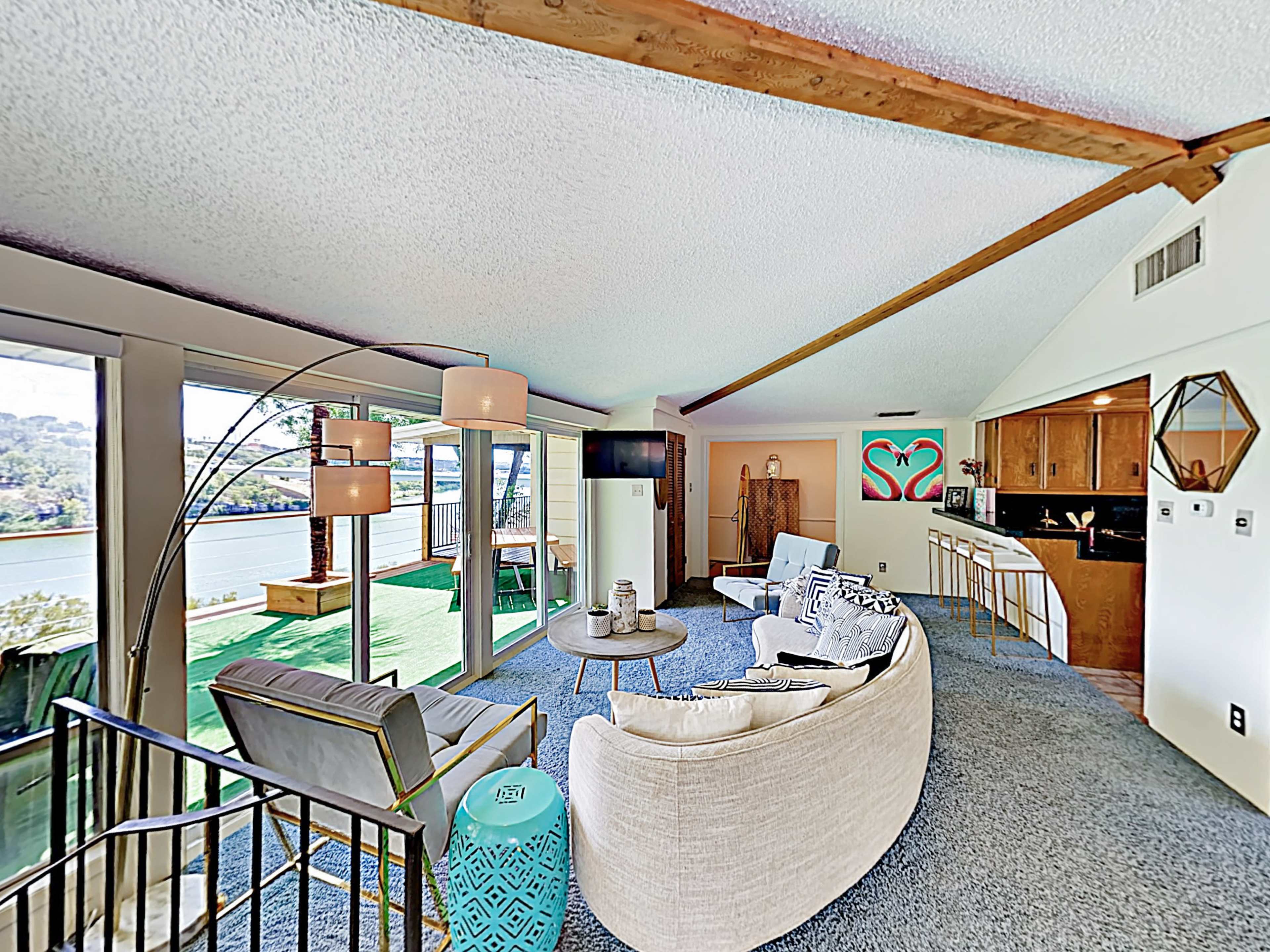 The image shows a well-lit living space with large windows overlooking a body of water, featuring modern furniture, a round table, and a kitchen area in the background.