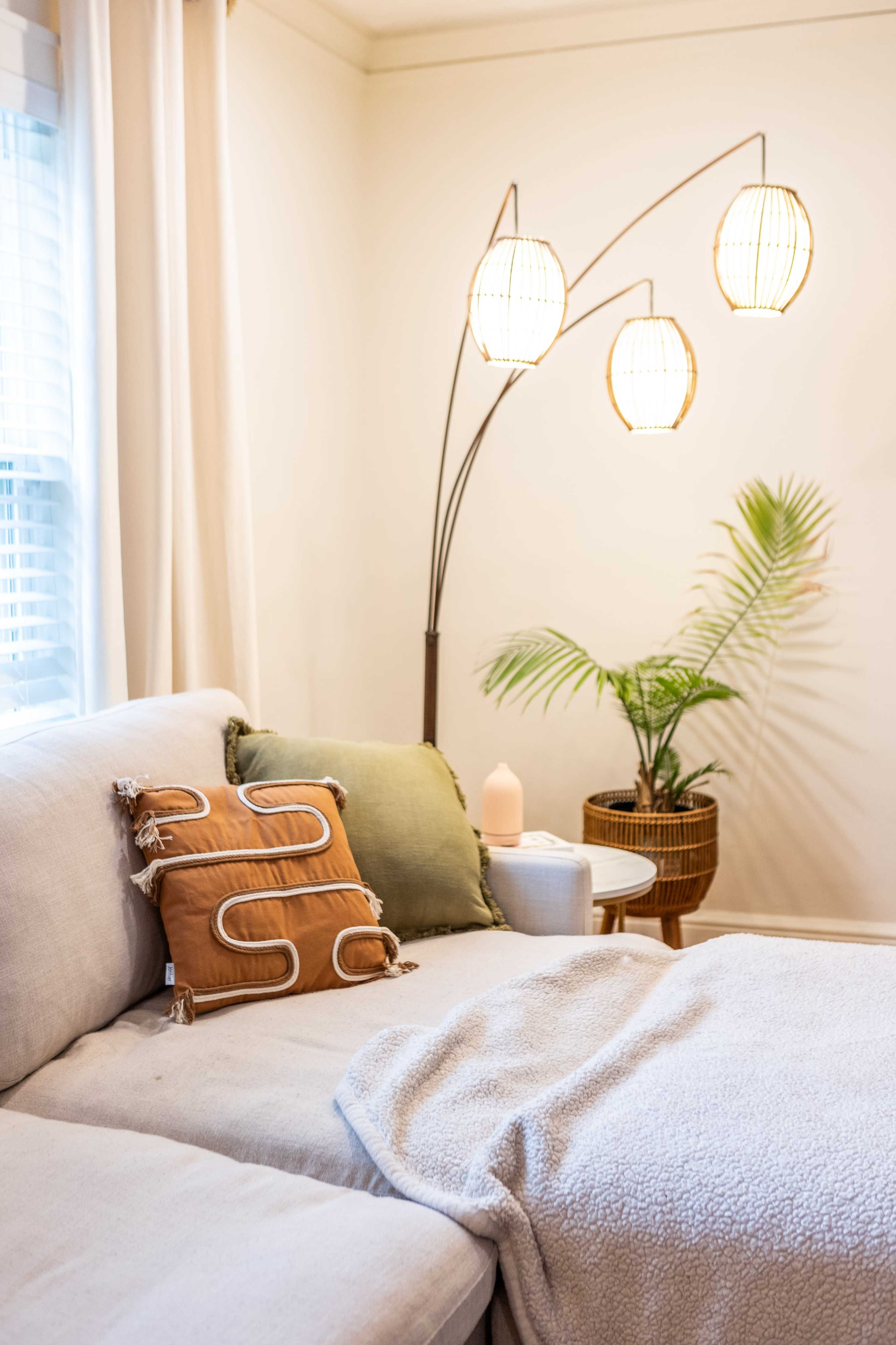 A cozy living room corner features a light-colored couch with decorative pillows, a small side table, and a floor lamp with three woven shades next to a potted plant.