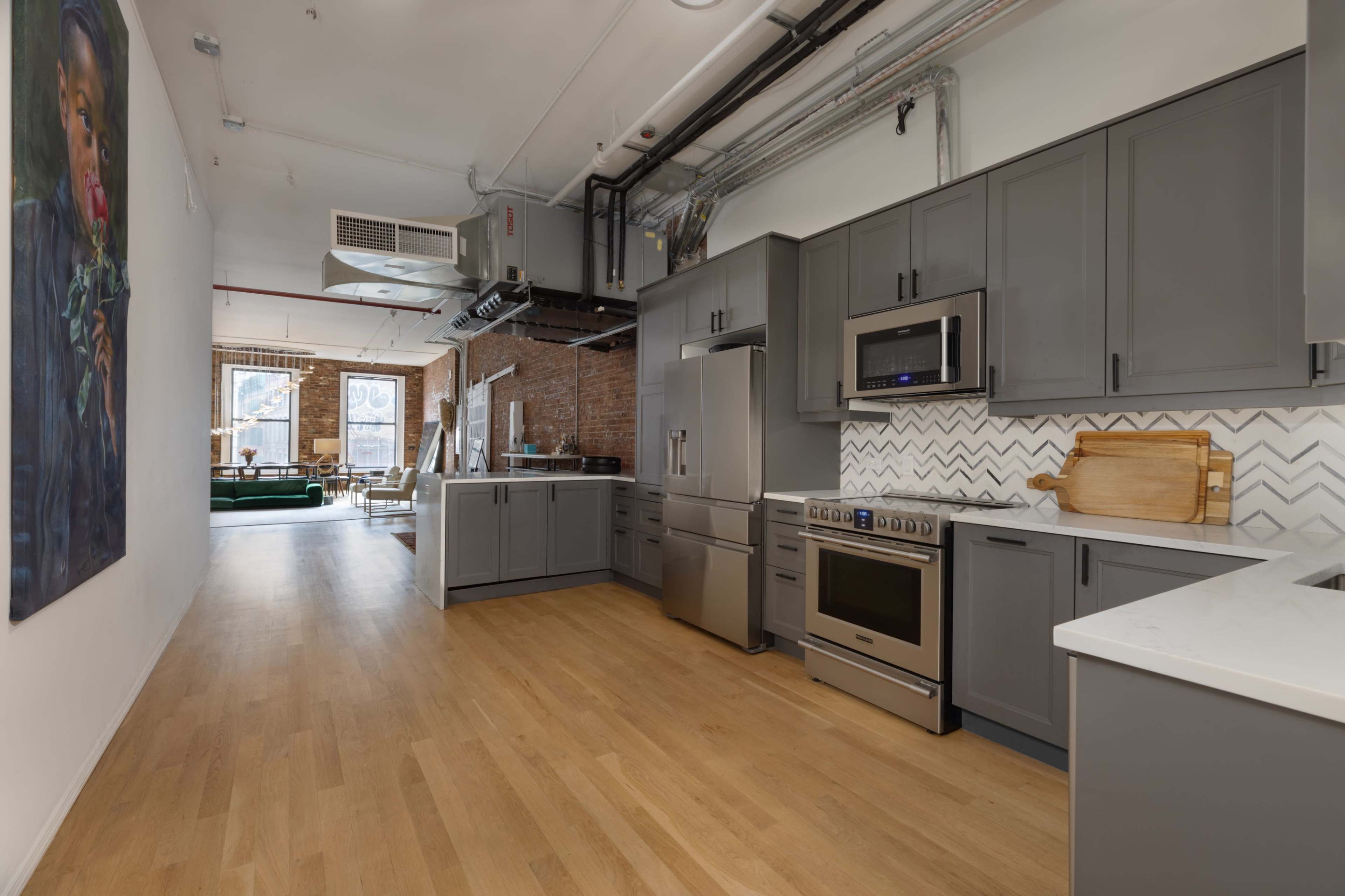 A modern kitchen with gray cabinets, stainless steel appliances, and a white countertop, adjacent to a bright open living space featuring exposed brick walls and large windows.