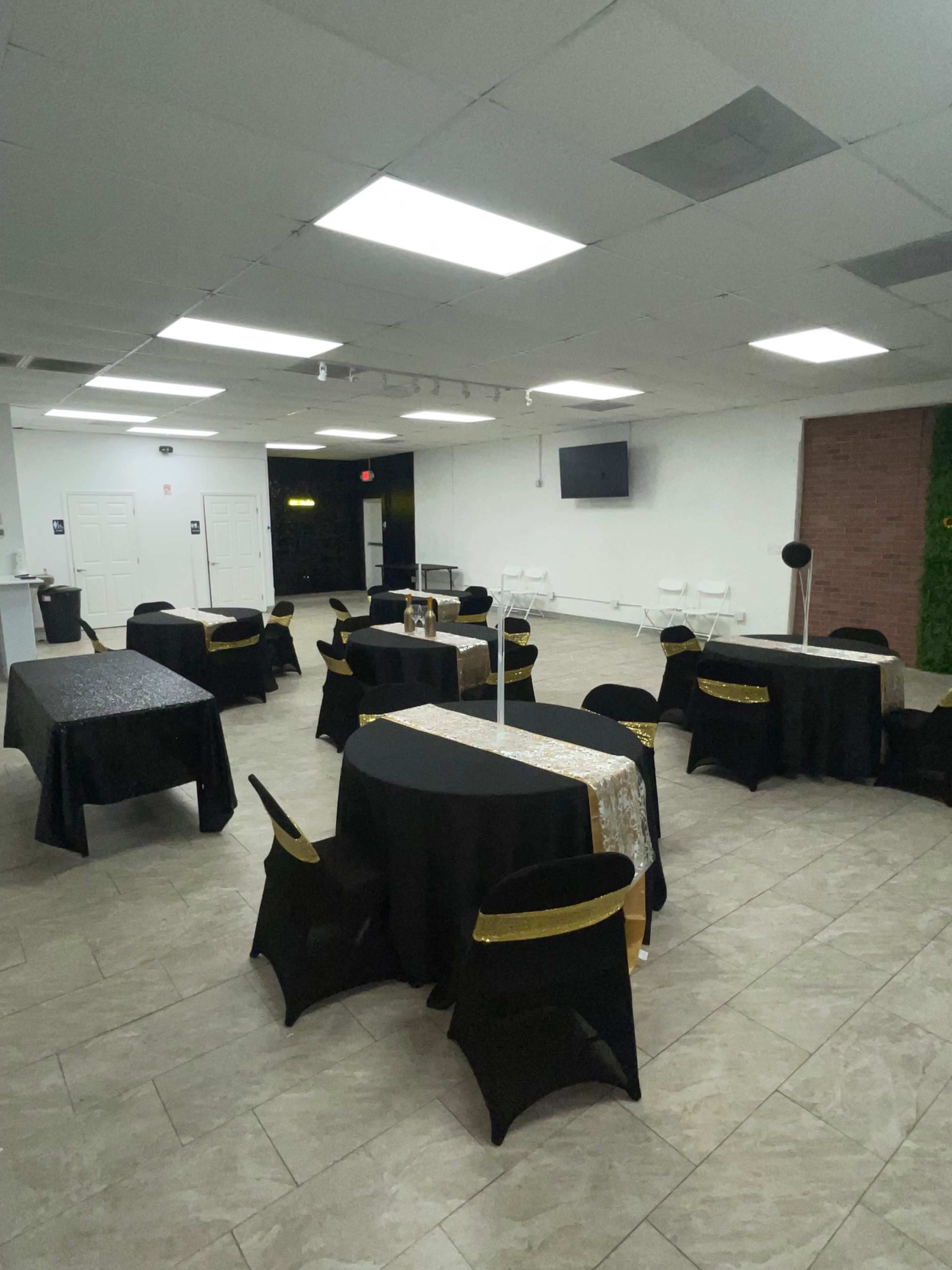 A room set up for an event, featuring tables covered in black and gold tablecloths arranged throughout the space with white chairs nearby.