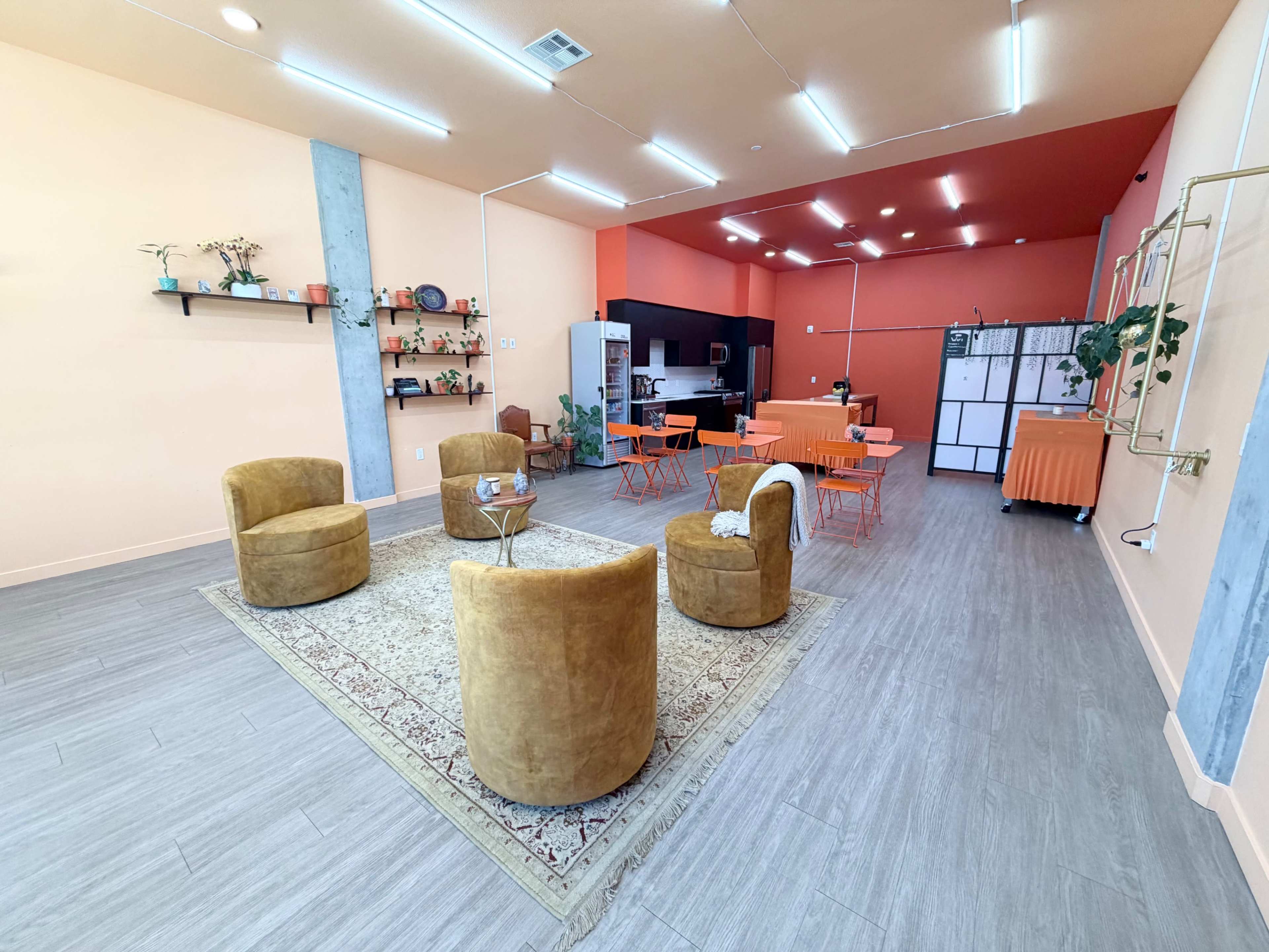 The image shows a modern, brightly colored room with orange and peach walls, featuring four round tan chairs arranged around a rug, a dining area with orange tables, and a kitchenette in the background.