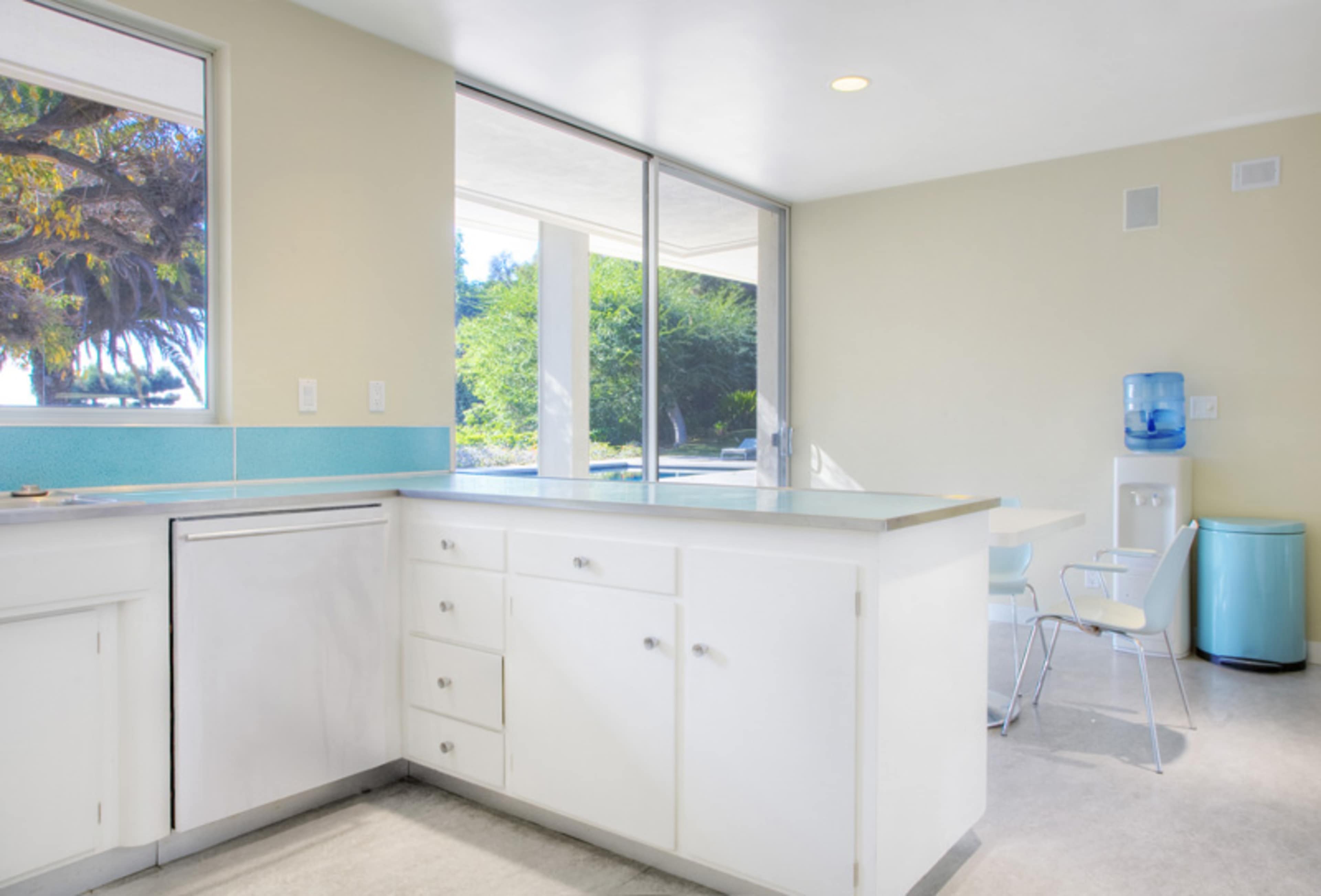 A bright kitchen with white cabinetry, a blue accent wall, a water cooler, and a dining table near large windows overlooking greenery.