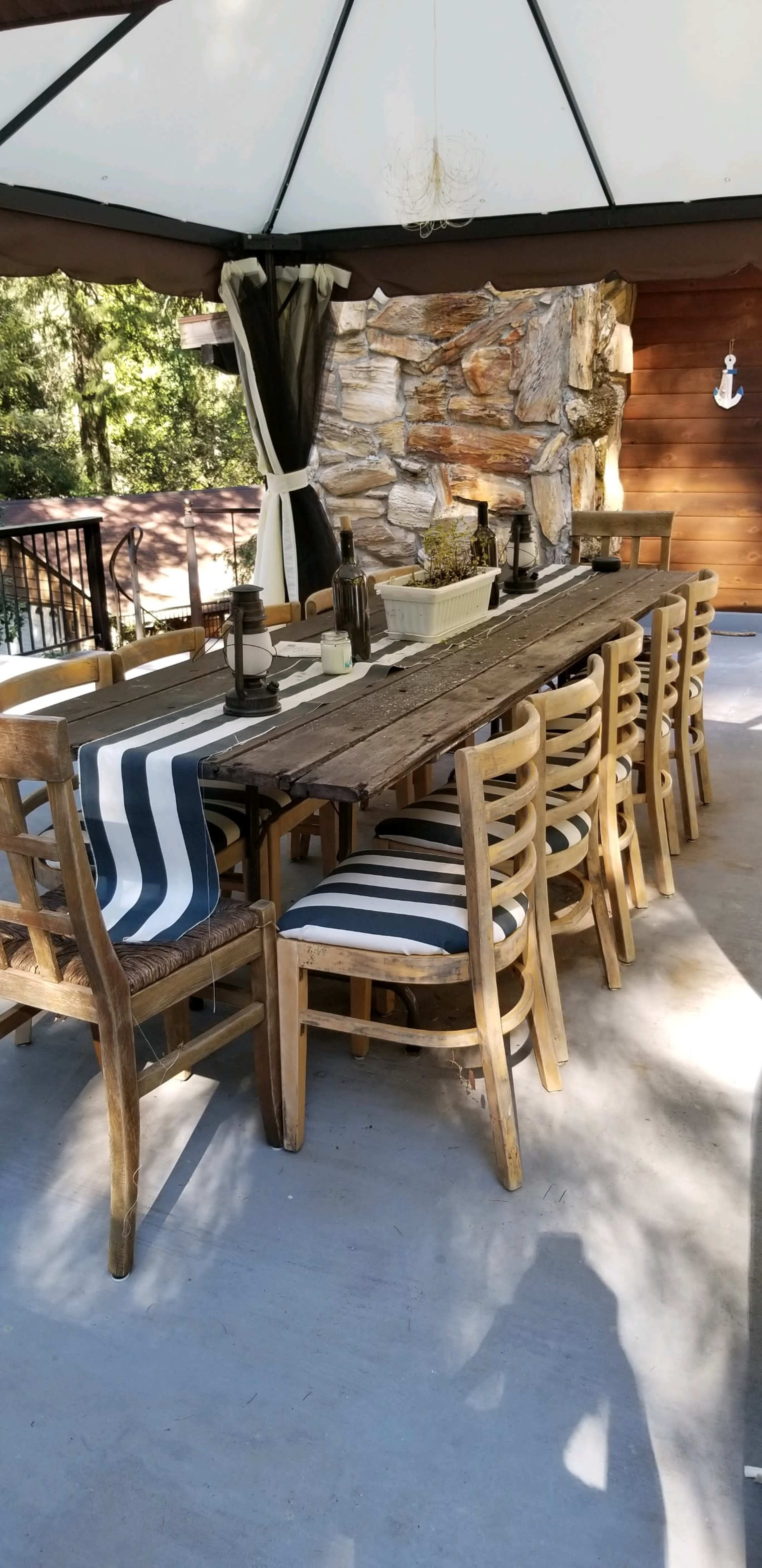 A long wooden table with striped chairs is set under a gazebo next to a stone fireplace.
