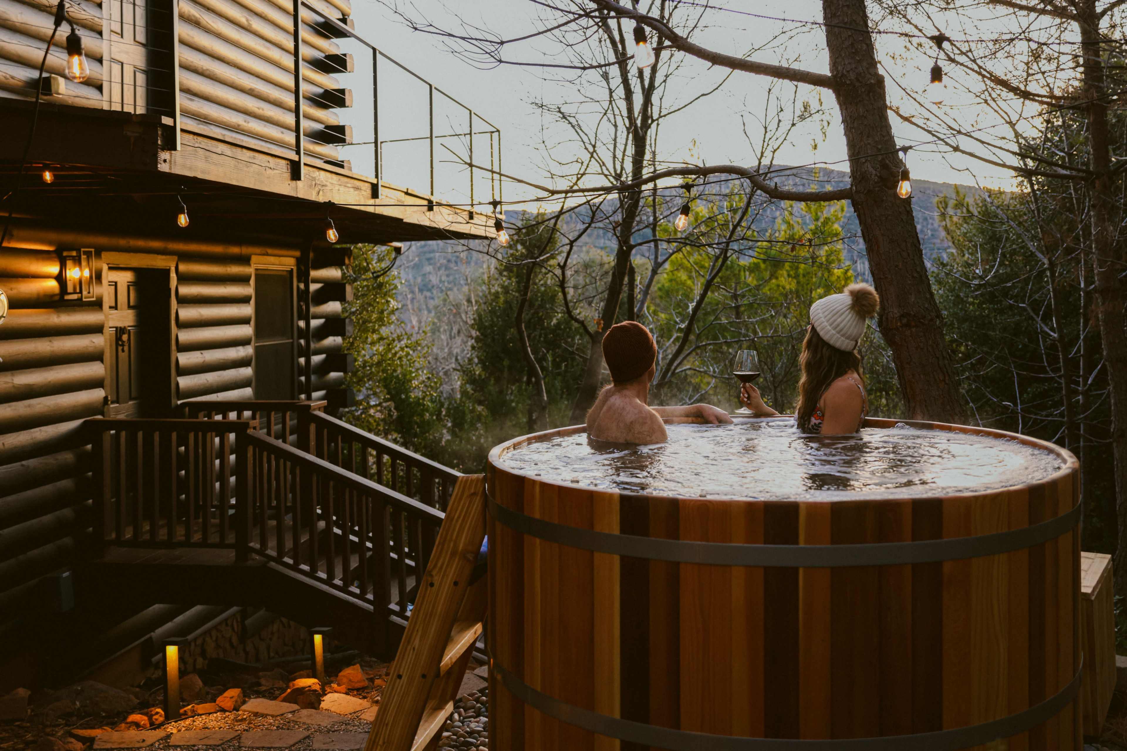 Two people relax in a wooden hot tub beside a cabin, surrounded by trees and mountains, while holding drinks.