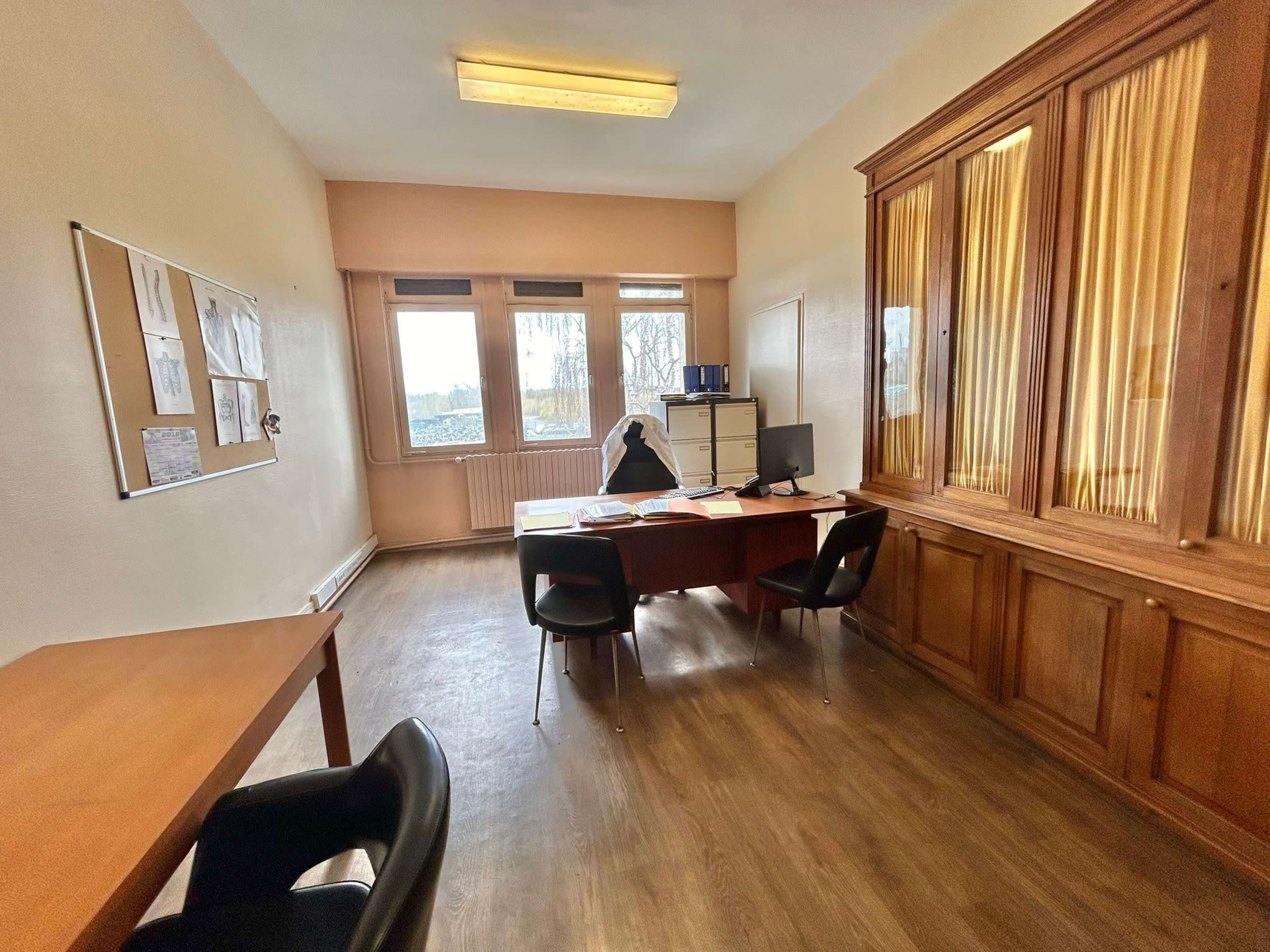 A simple office space with a wooden desk, black chairs, a computer, and large windows letting in natural light.