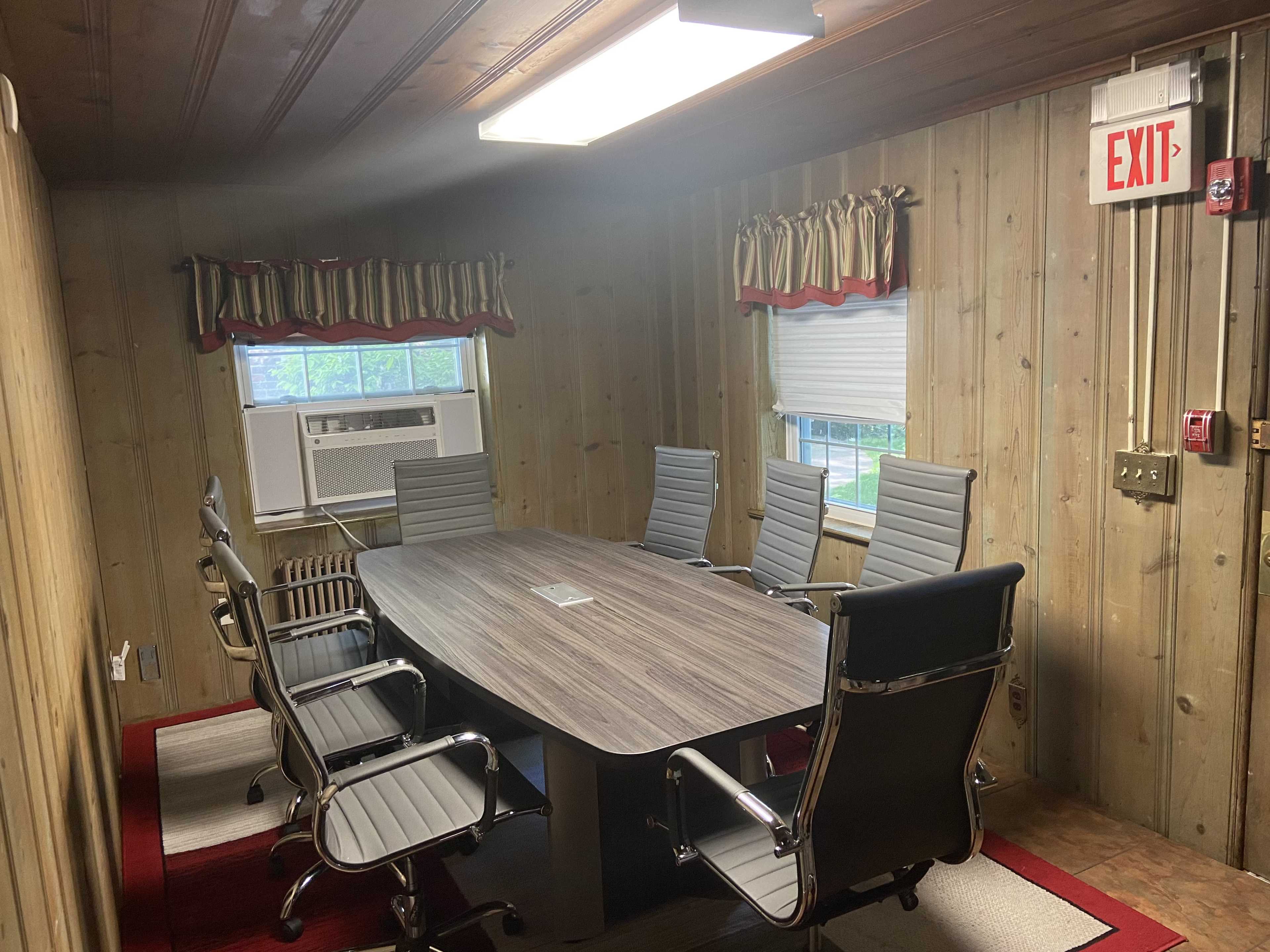 A conference room features a long oval table surrounded by six ergonomic chairs, with wooden paneling on the walls and an air conditioning unit visible in the corner.