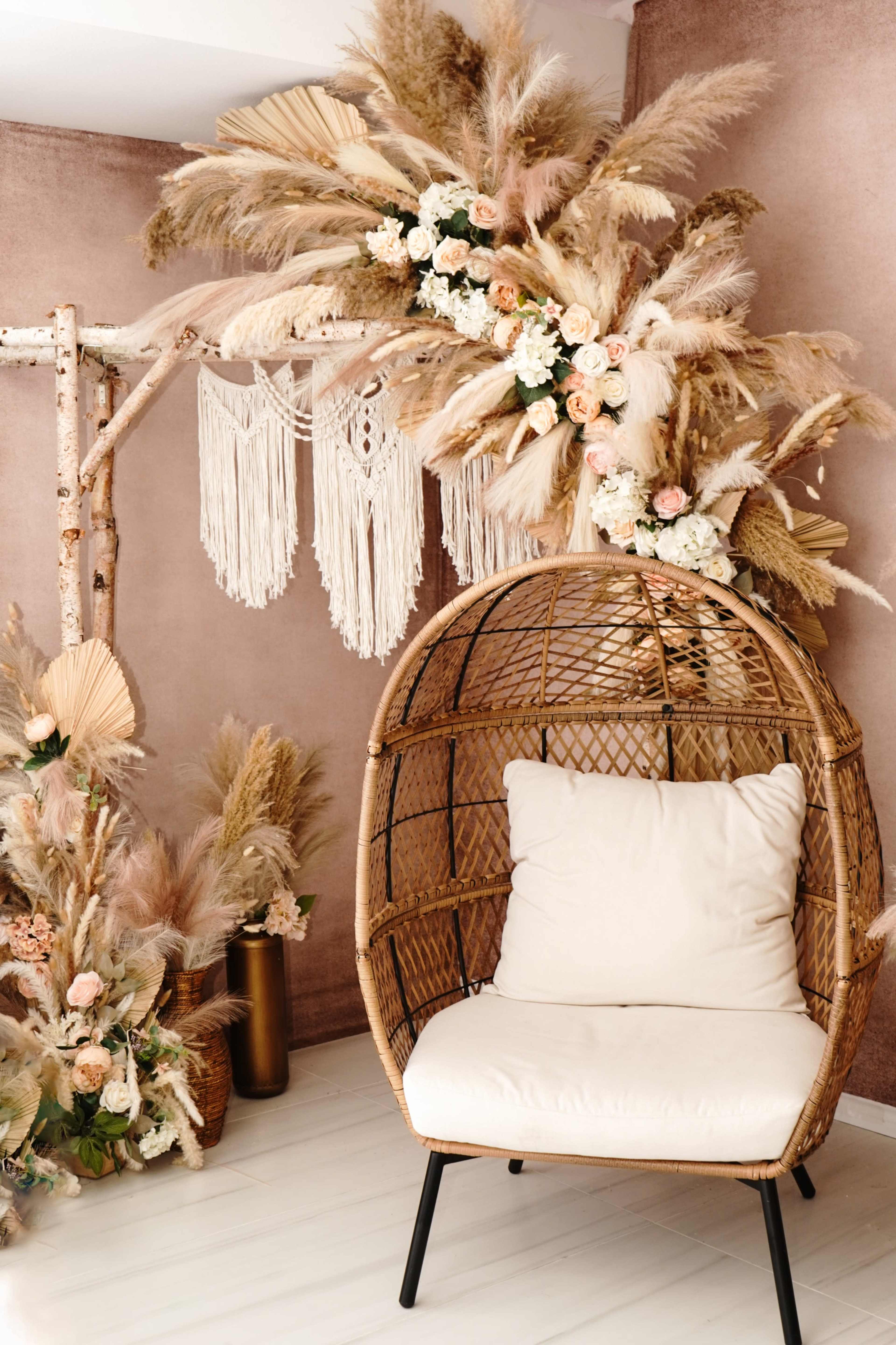 The image features a stylish sitting area with a wicker hanging chair, adorned with decorative flowers and pampas grass, set against a textured brown wall.