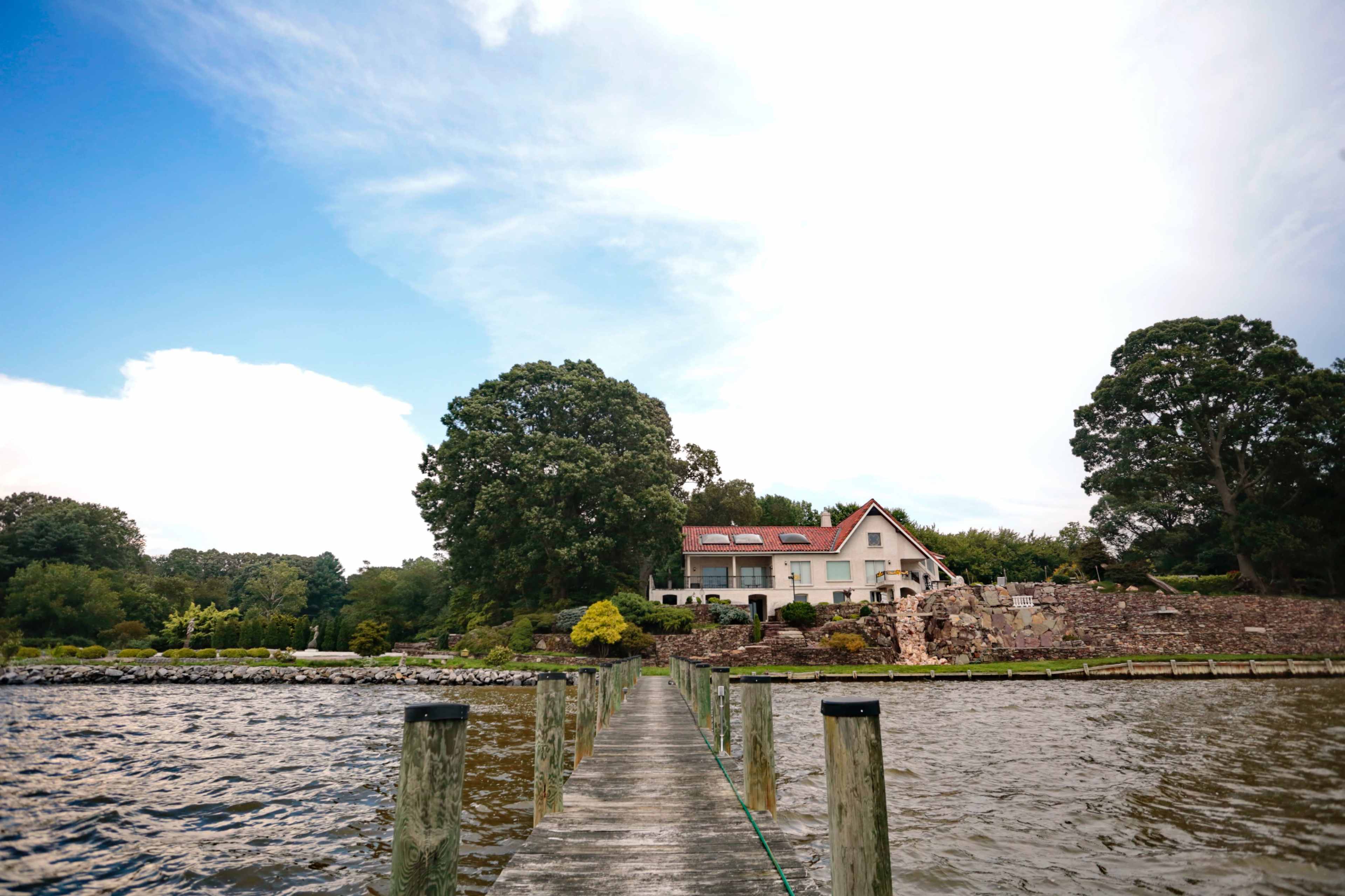 A wooden dock extends over the water toward a large house surrounded by greenery.