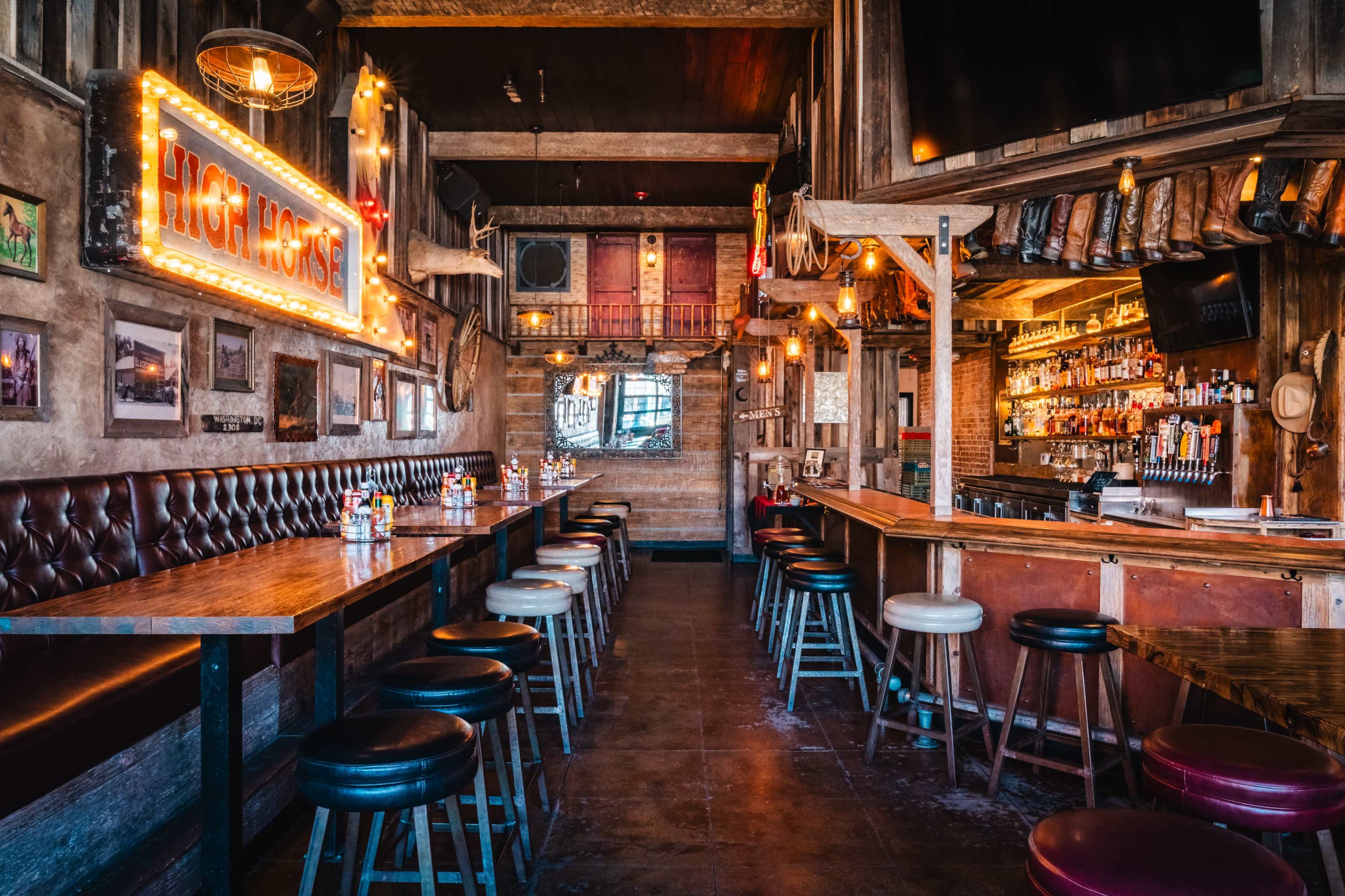 The image shows the interior of a rustic bar with wooden seating, a well-stocked bar, and bright neon signage.