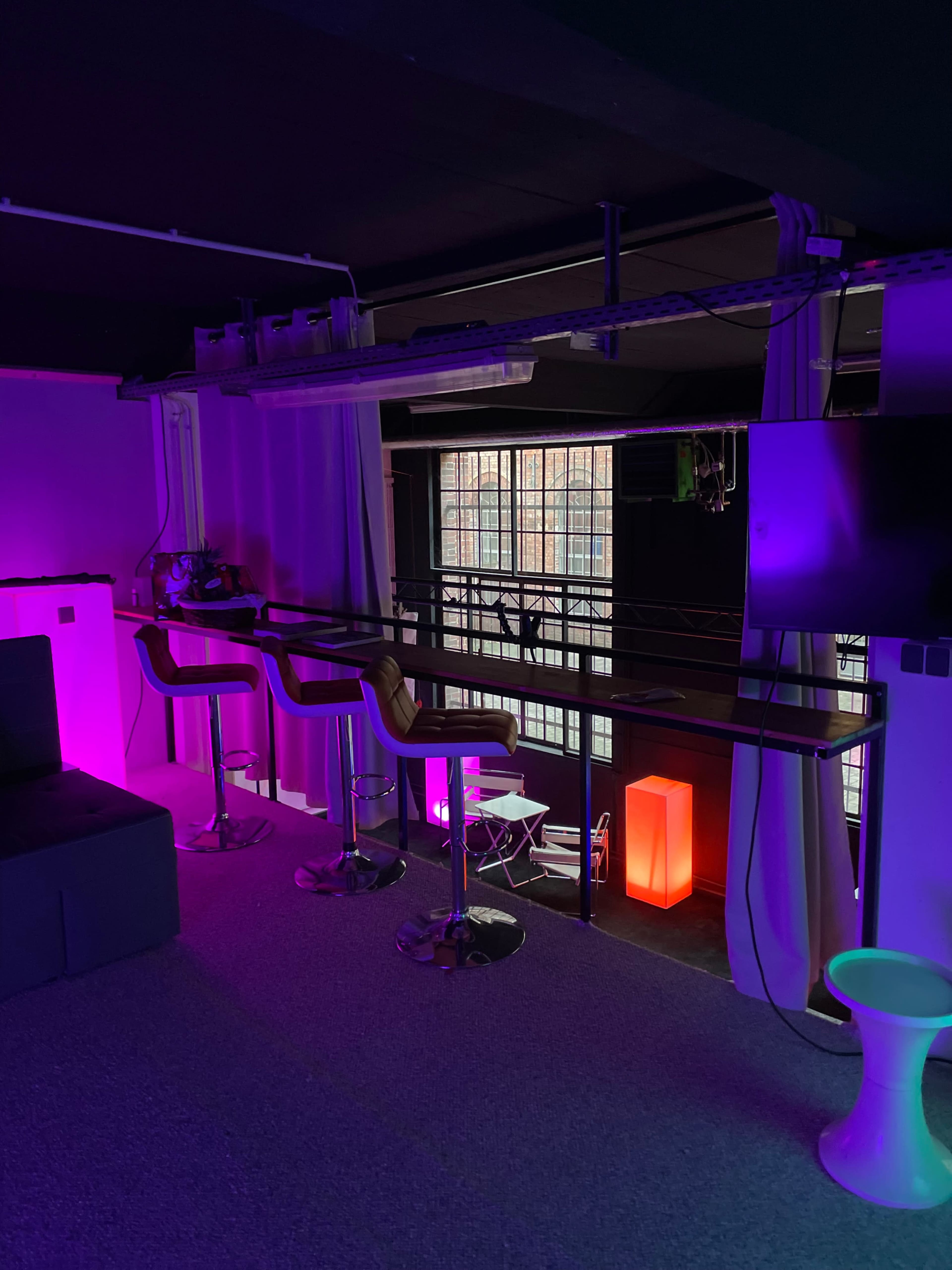 The image shows a dimly lit room featuring modern barstools by a counter, illuminated by purple and red lights, with large windows in the background.