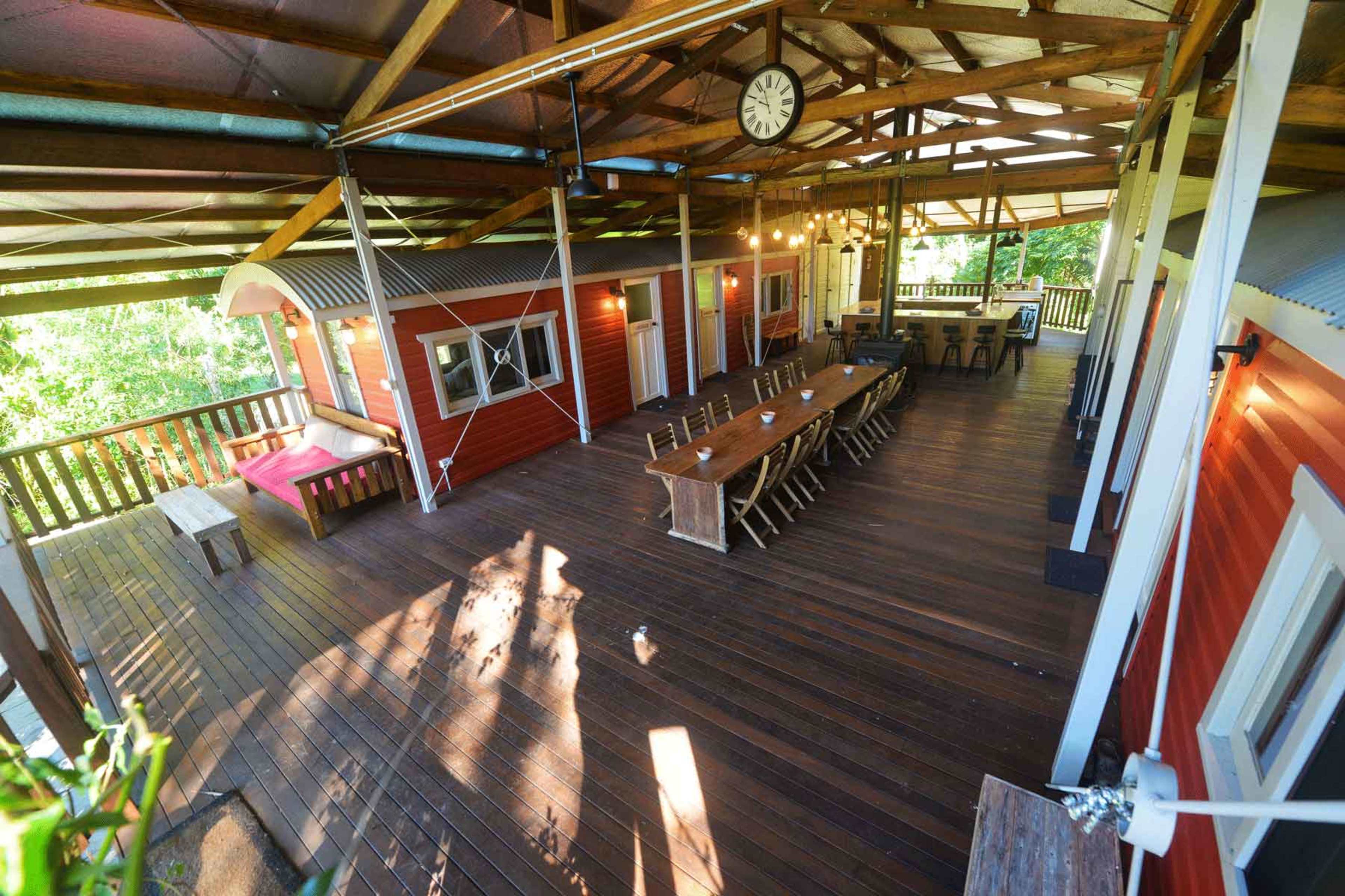 The image depicts a spacious wooden deck with several seating areas, including a long dining table and a smaller seating arrangement, all beneath a covered structure supported by wooden beams.