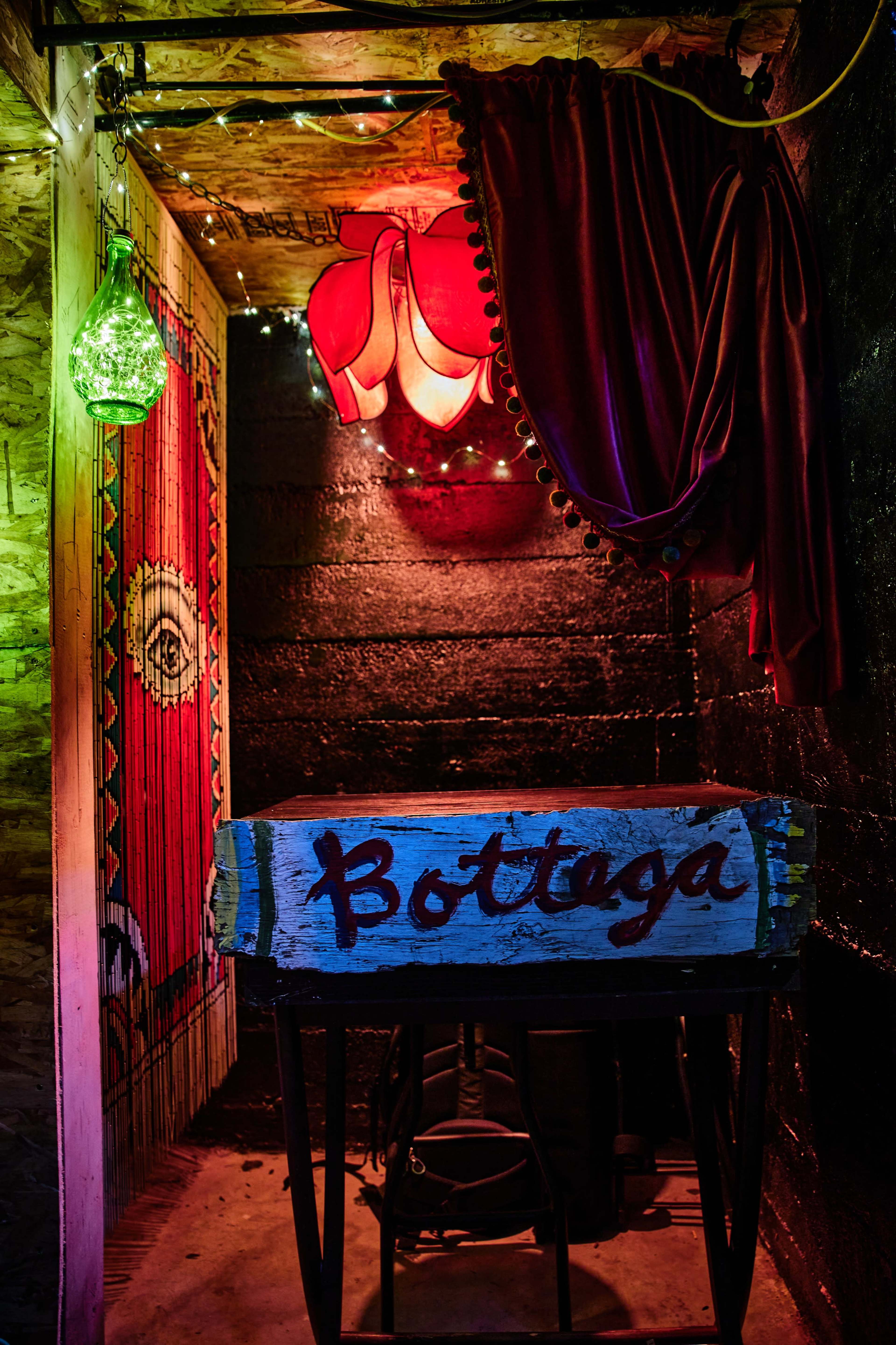 A dimly lit corner of a room with a wooden table labeled "Bottega," colorful lighting, and decorative fabric hanging nearby.
