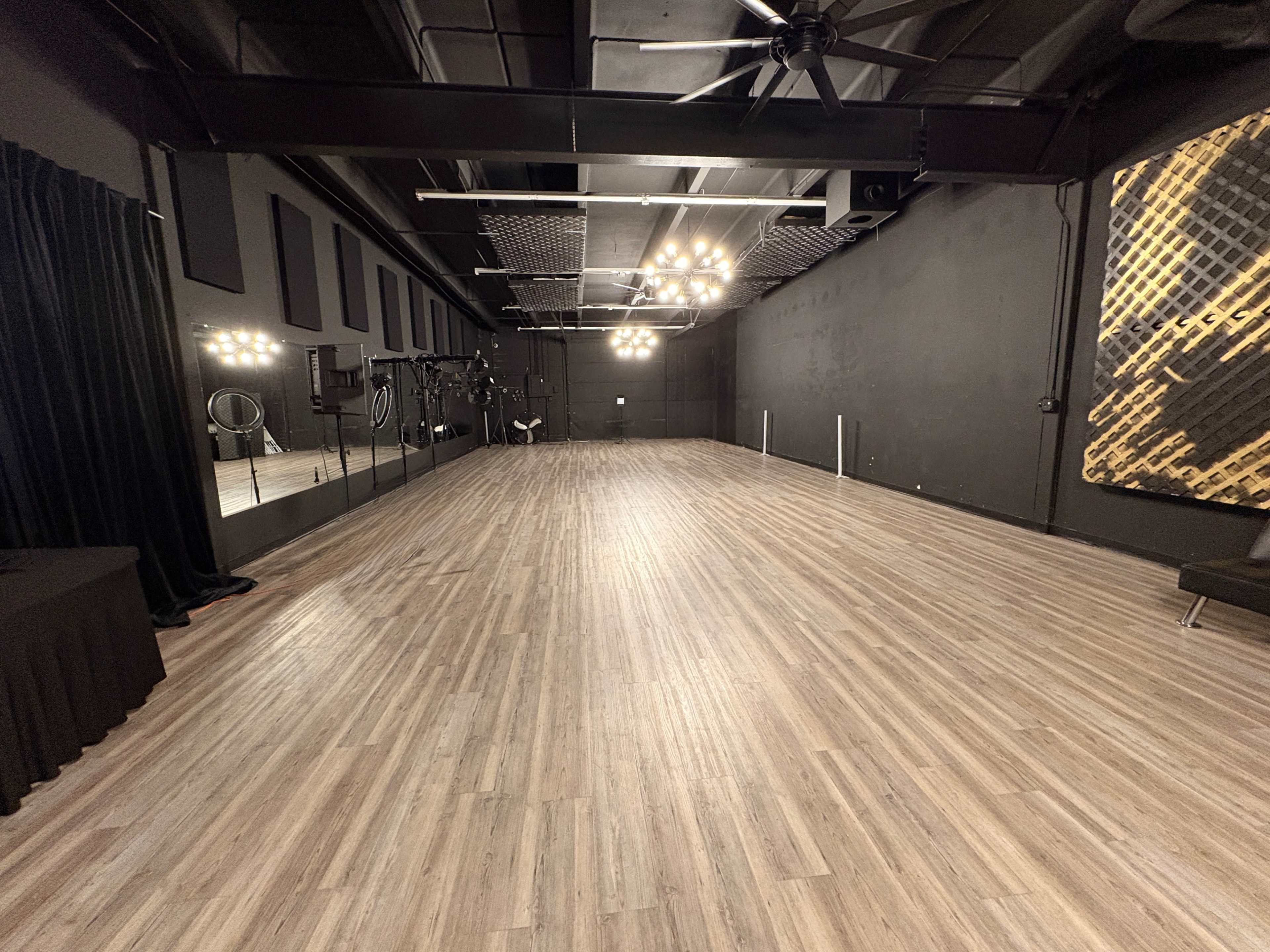 The image shows a spacious room with wooden flooring, black walls, large mirrors, and overhead lighting fixtures.