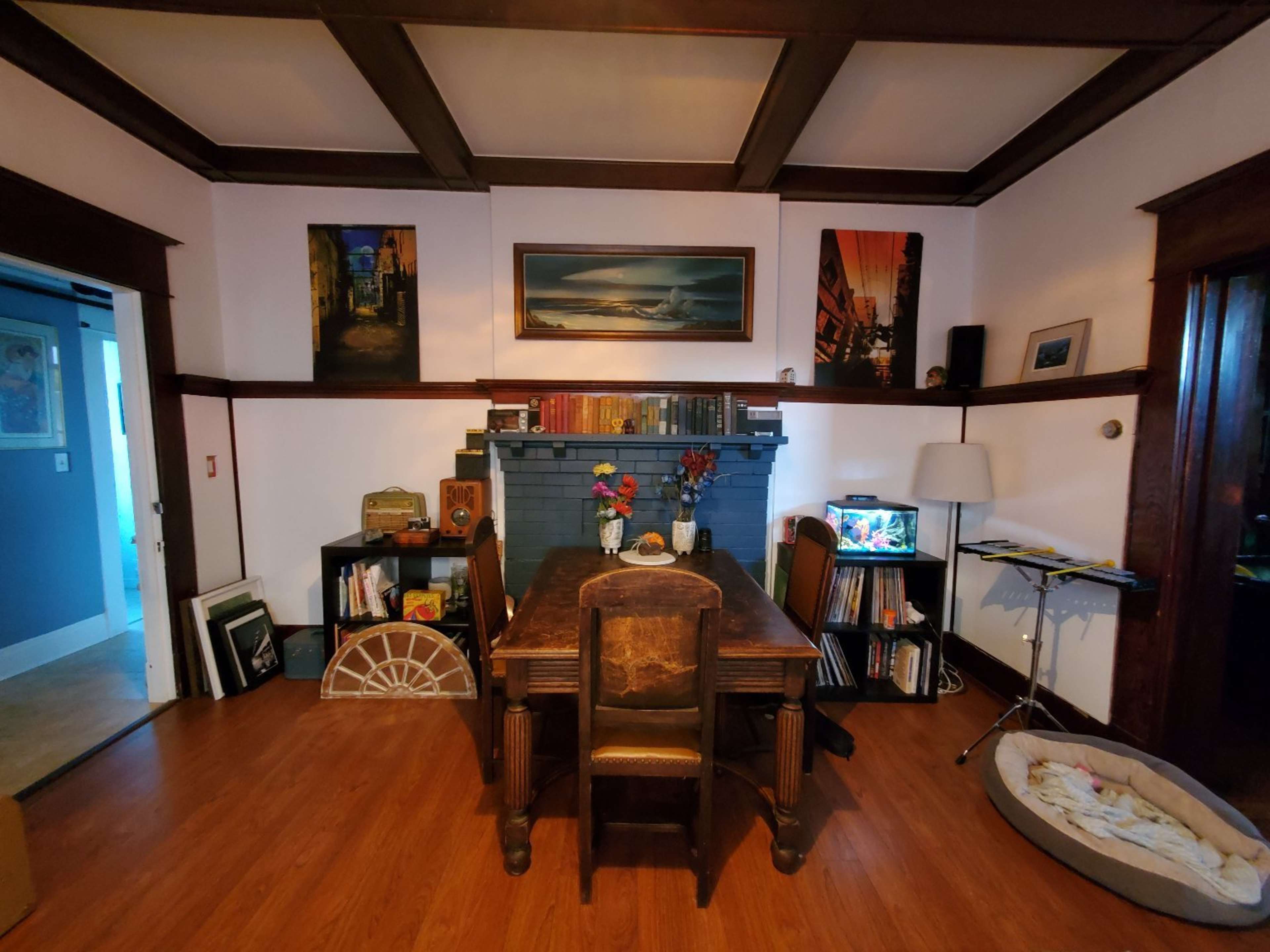 A cozy dining area with a wooden table surrounded by chairs, a blue-painted fireplace, and shelves filled with books and decorative items.