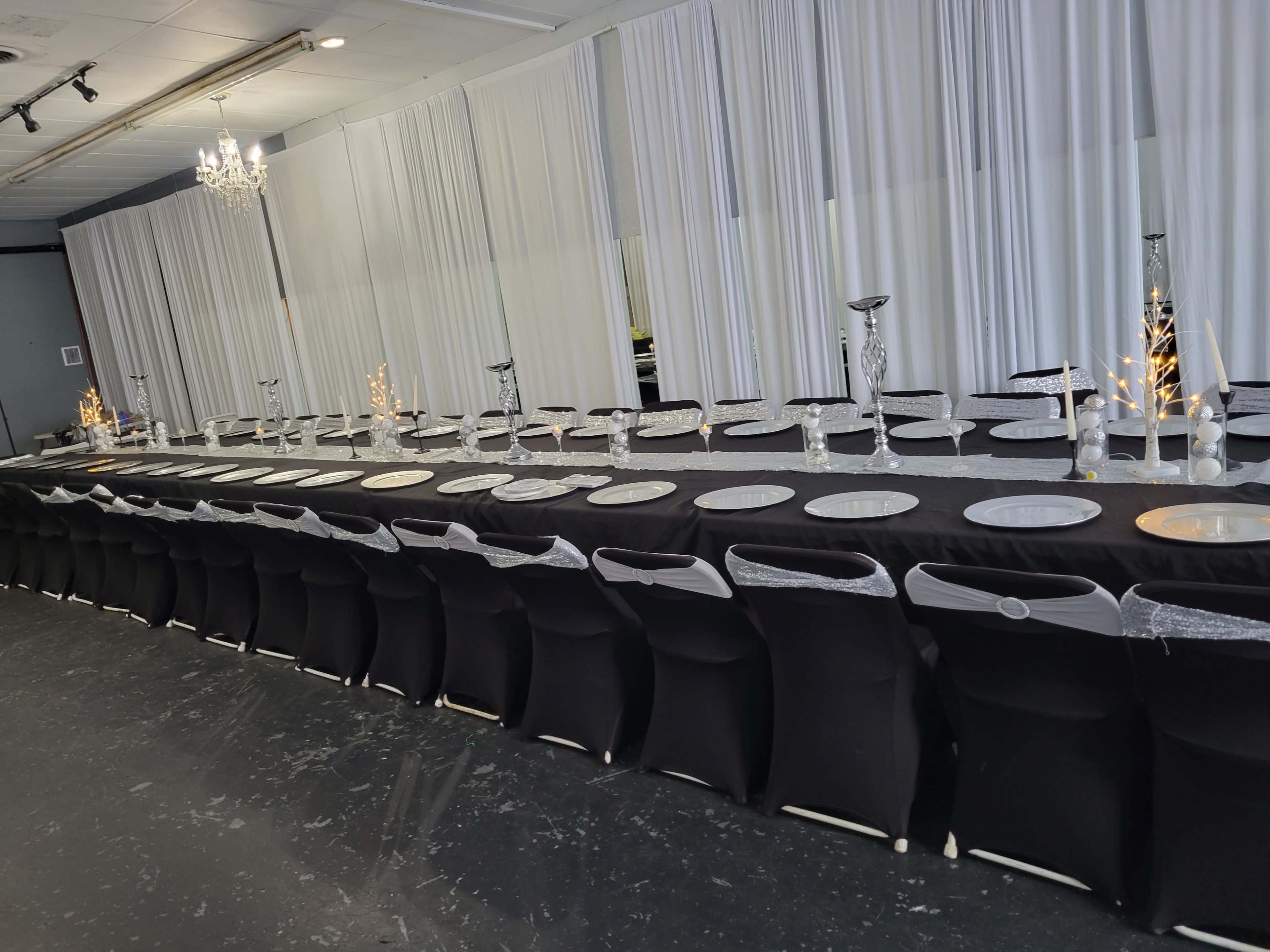 A long dining table with black tablecloths and white plates is set up in a room with white drapes and a chandelier.