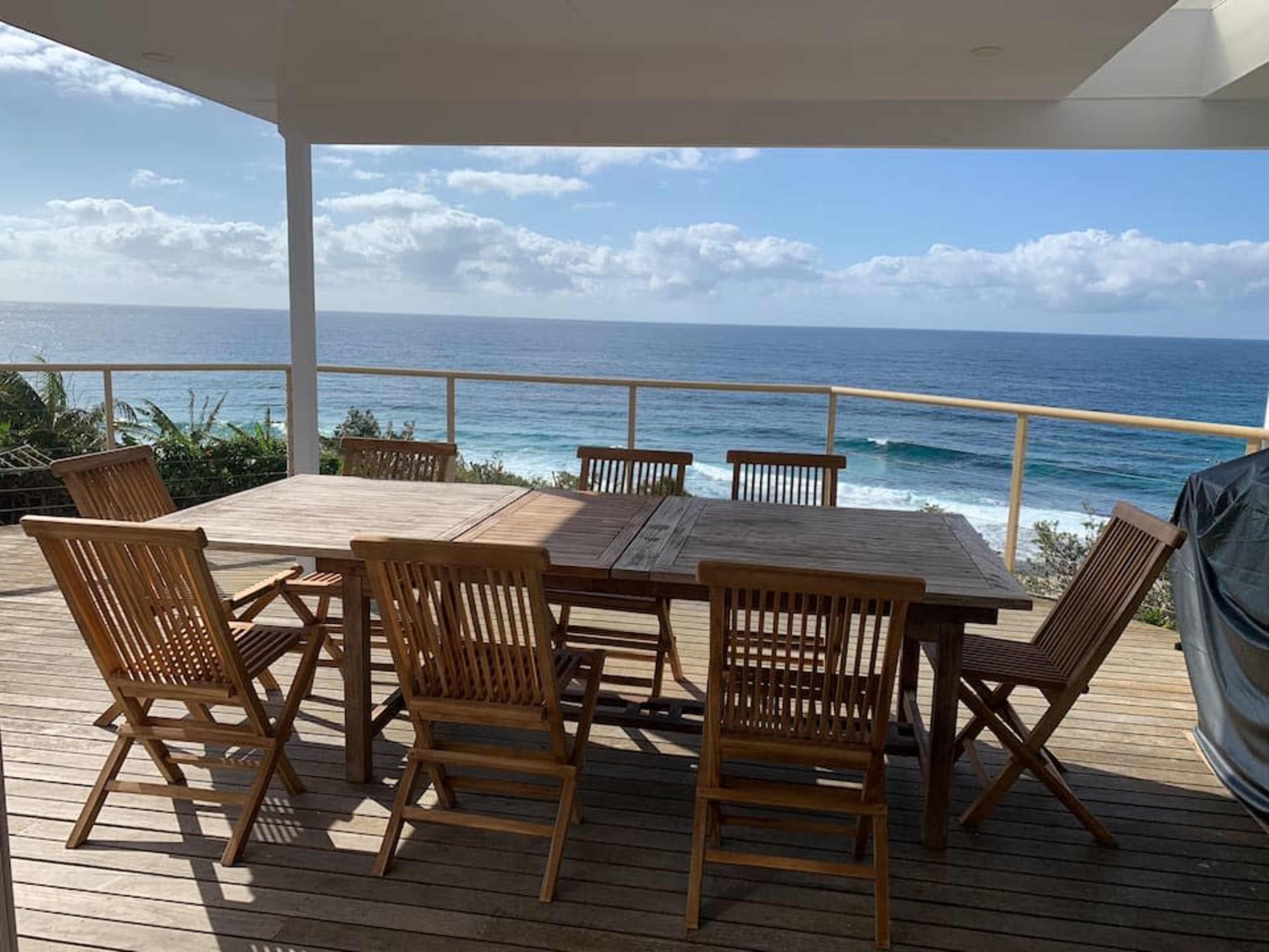 A wooden dining table with eight chairs is situated on a deck overlooking the ocean.
