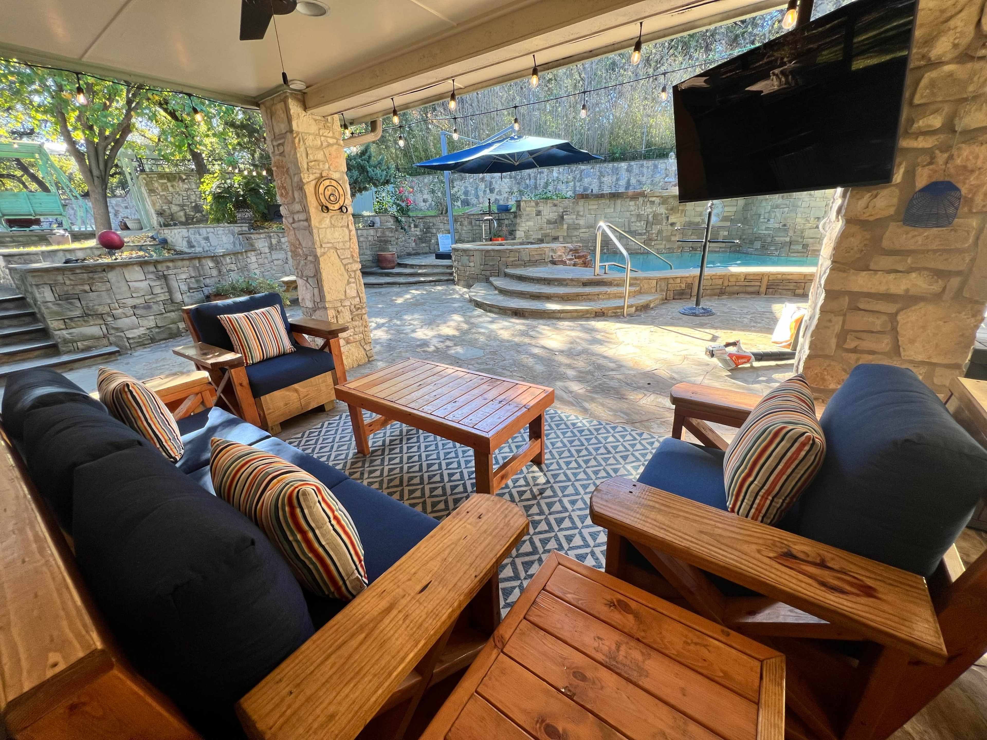 The image shows a patio area with wooden furniture, including a sofa and coffee table, overlooking a pool and garden space.