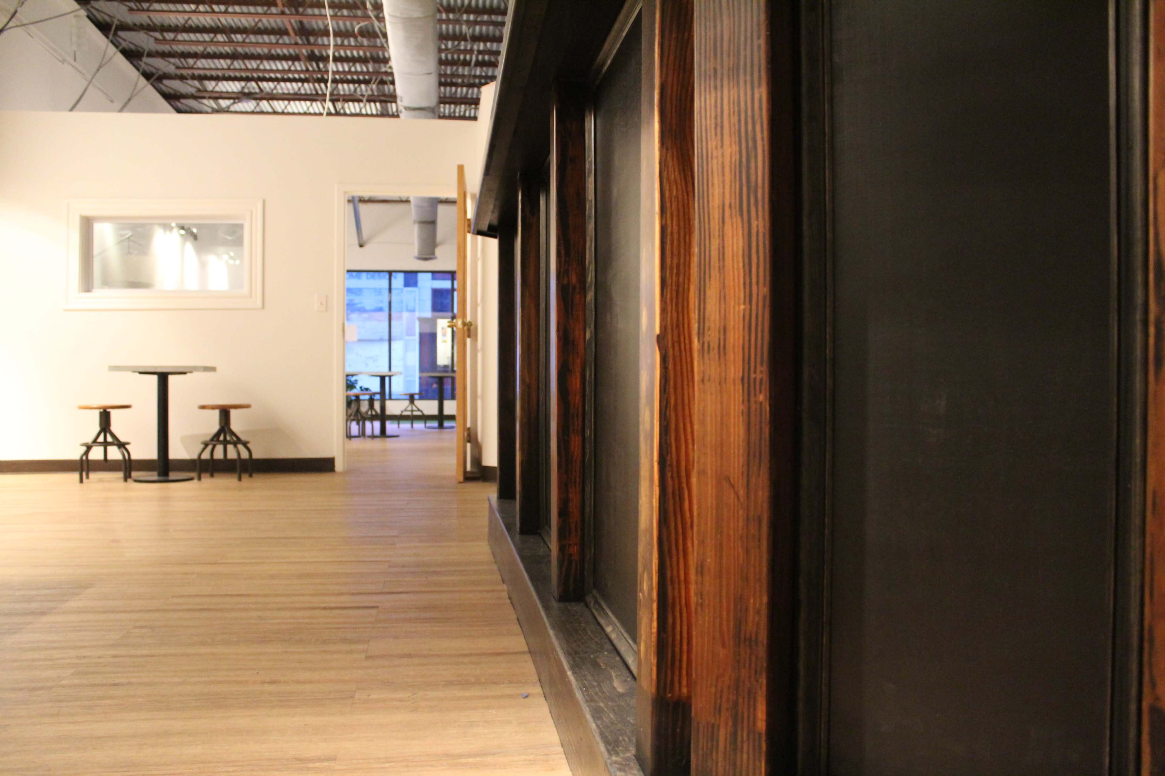 A modern interior space with wooden flooring, walls, and a table with two stools in the foreground, leading to an open area in the background.