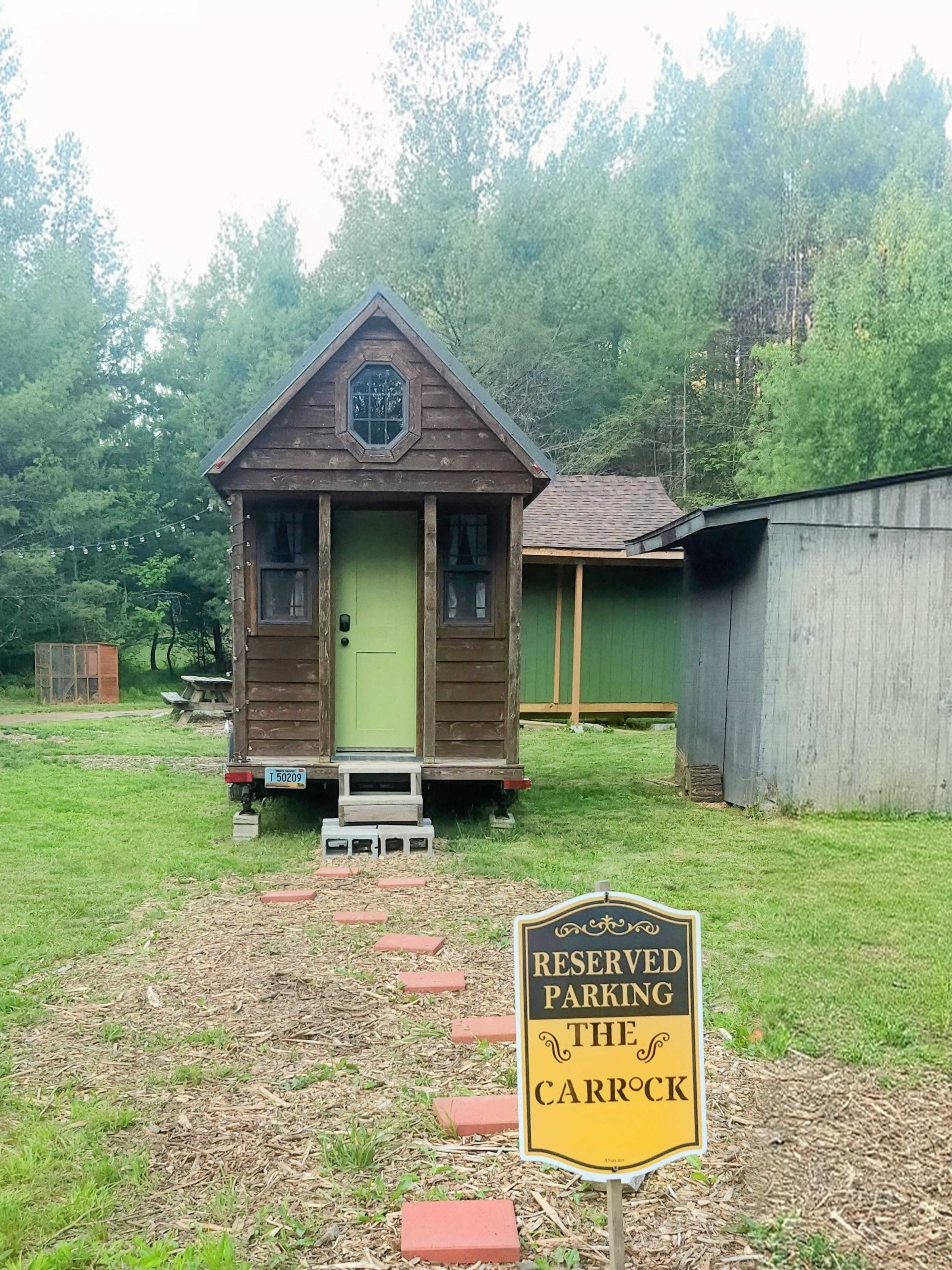The image shows a small, wooden cabin with a green door set on a trailer, accompanied by a sign that reads "Reserved Parking The Carrock," amidst a grassy area surrounded by trees.