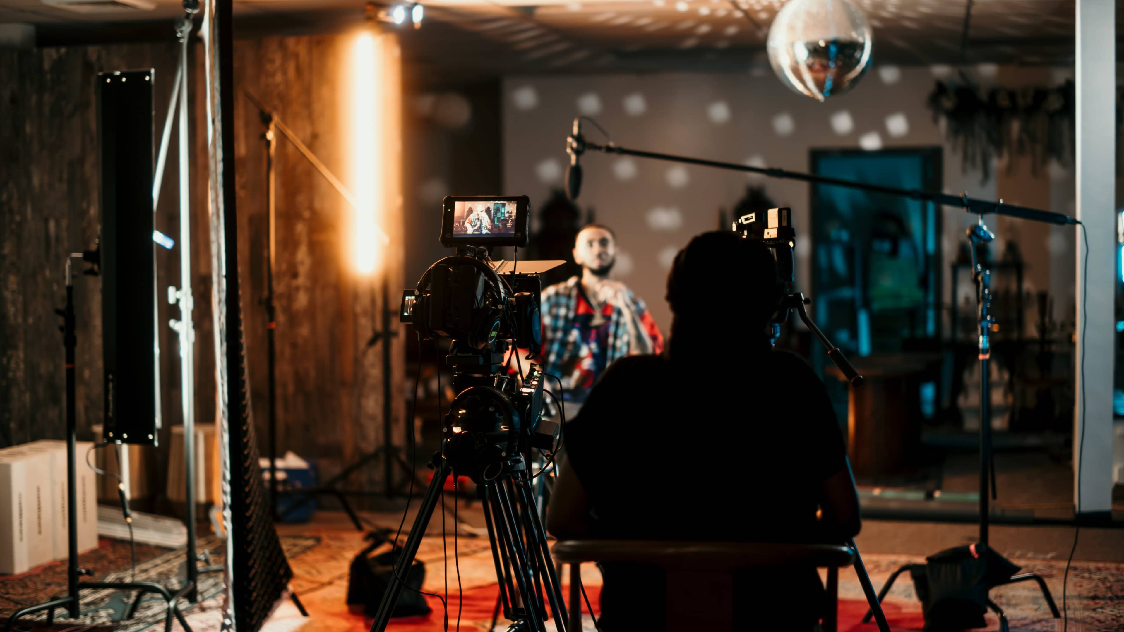A person sits in a chair in a studio setting with cameras and lighting equipment aimed at another person who is speaking in front of a backdrop.