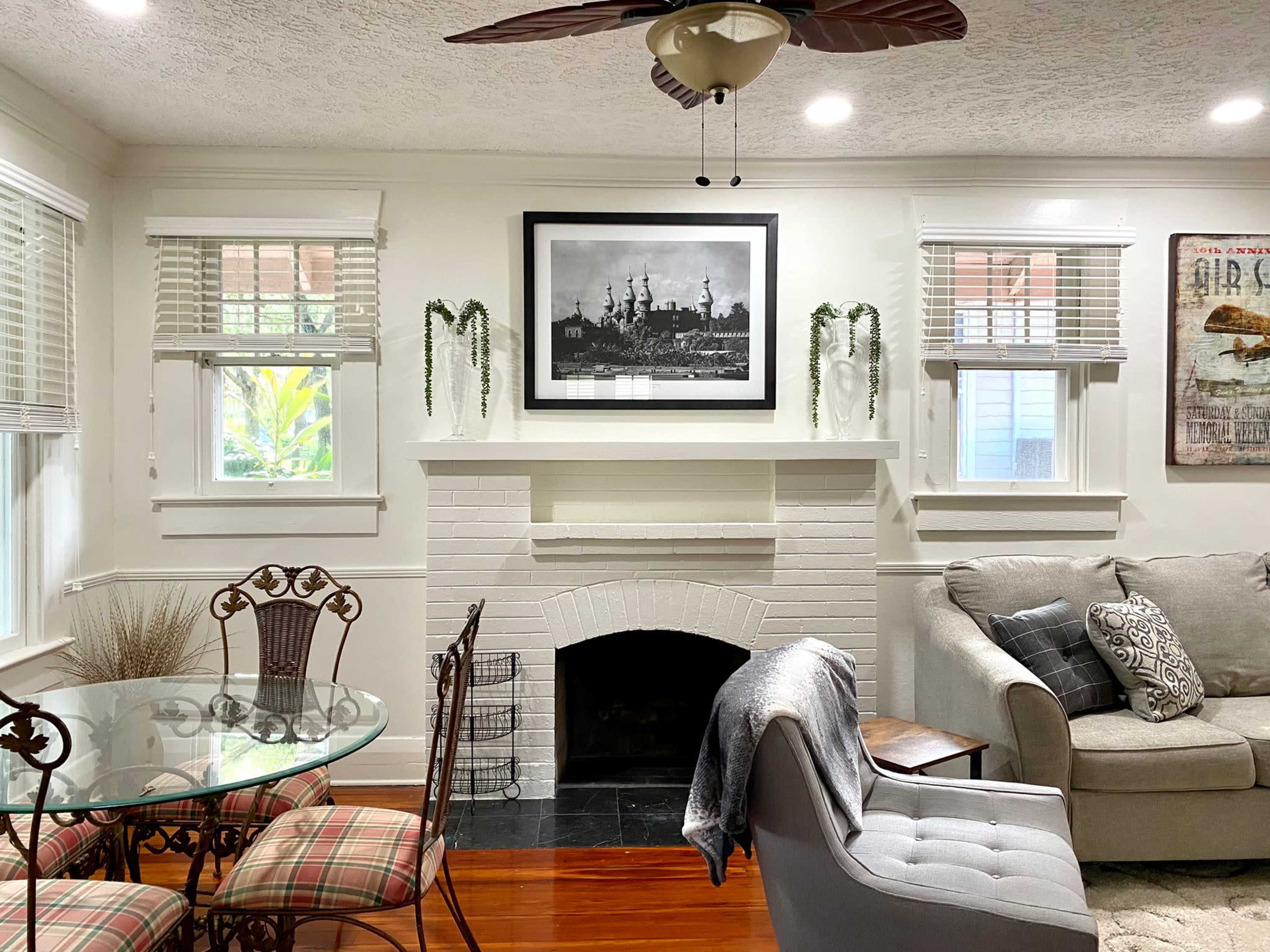 The image shows a living room featuring a white brick fireplace, a dining table with chairs, a gray sofa, and a large black-and-white photograph on the wall.
