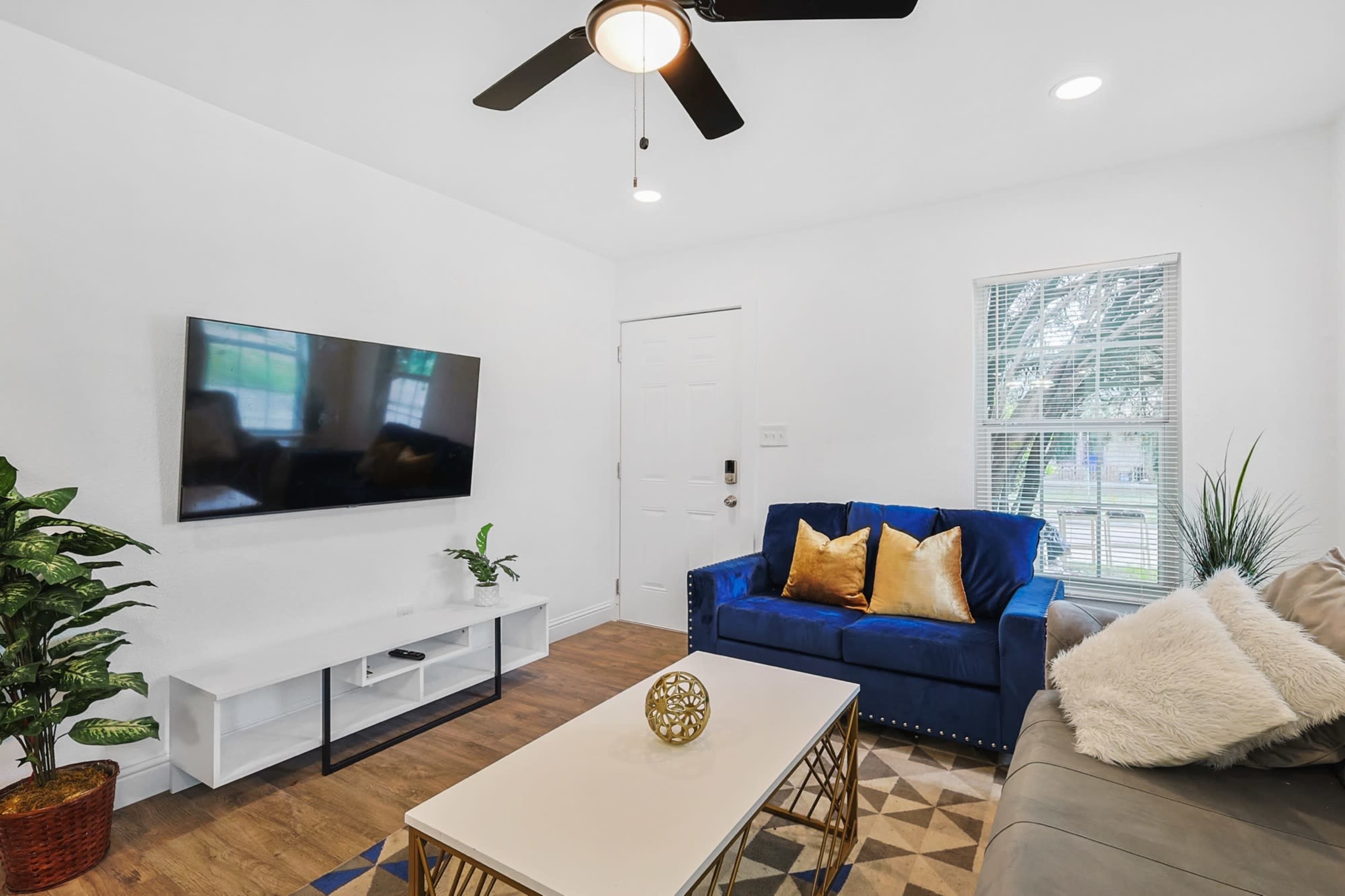 A living room features a blue sofa, a light-colored coffee table, a mounted television, and plants.