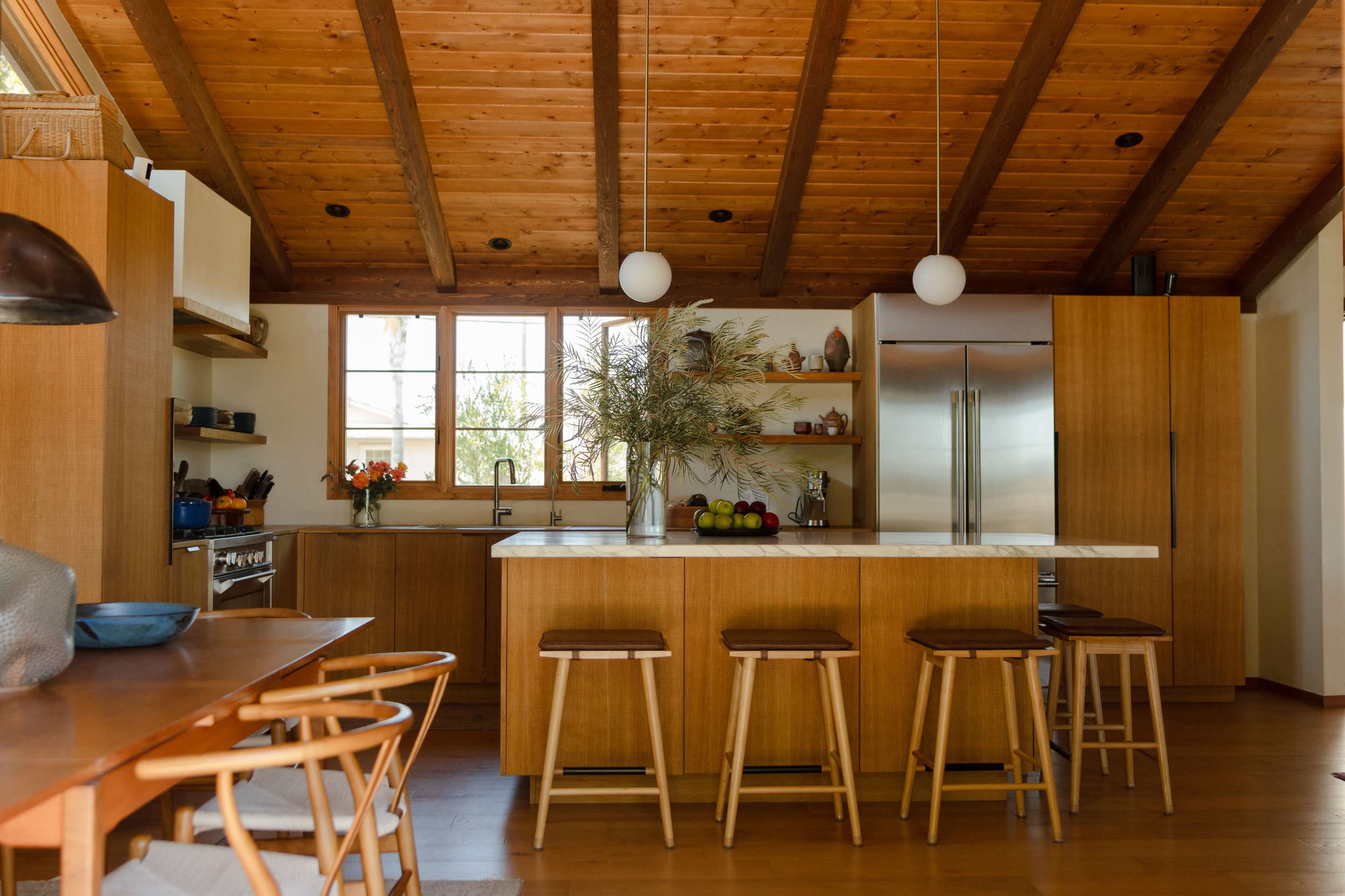The image shows a modern kitchen featuring wooden cabinets, a large island with bar stools, and ample natural light from the windows.