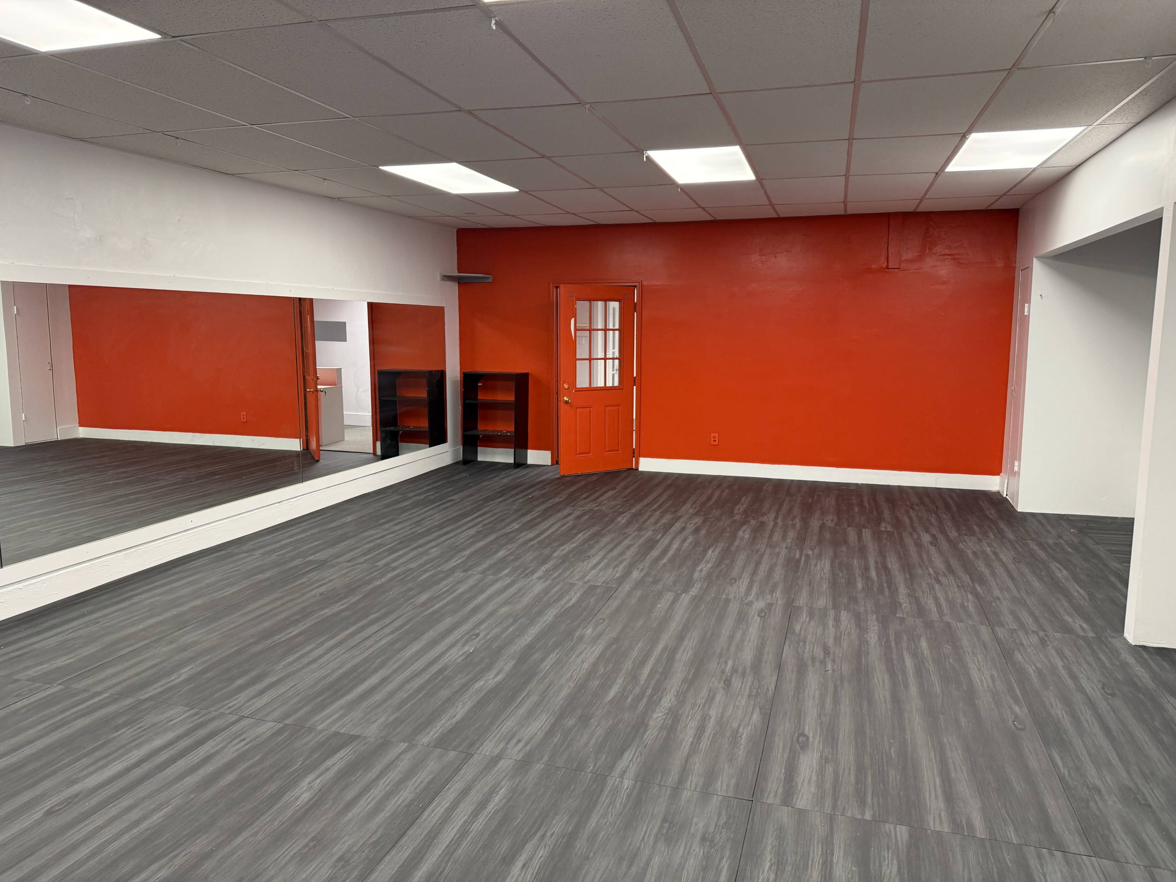 The image shows a spacious interior room with a red accent wall, a large mirror on one side, and a door leading to another area.