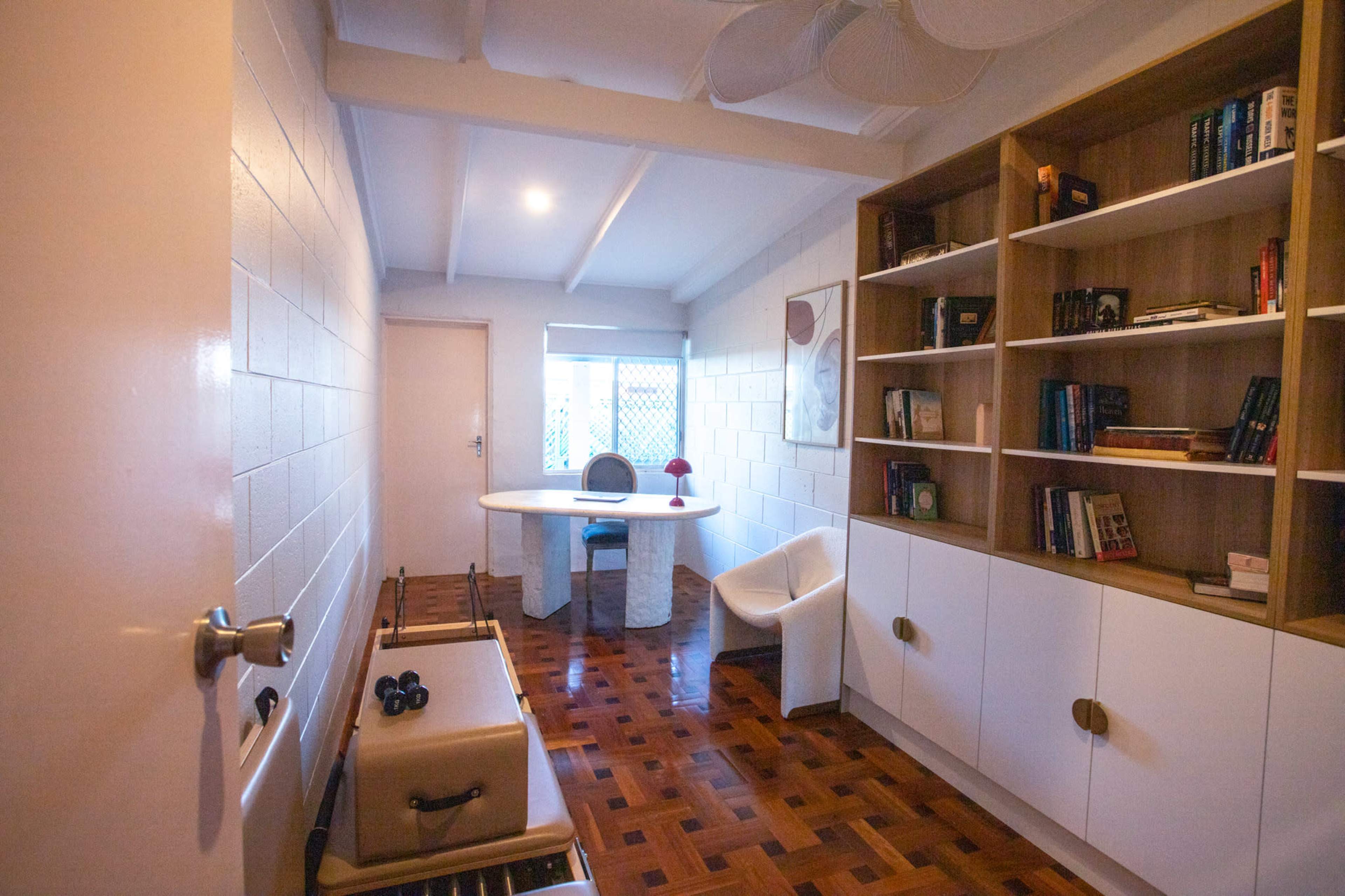 The image shows a minimalist room featuring a round table, shelving unit, and a chair, with wooden flooring and white walls.
