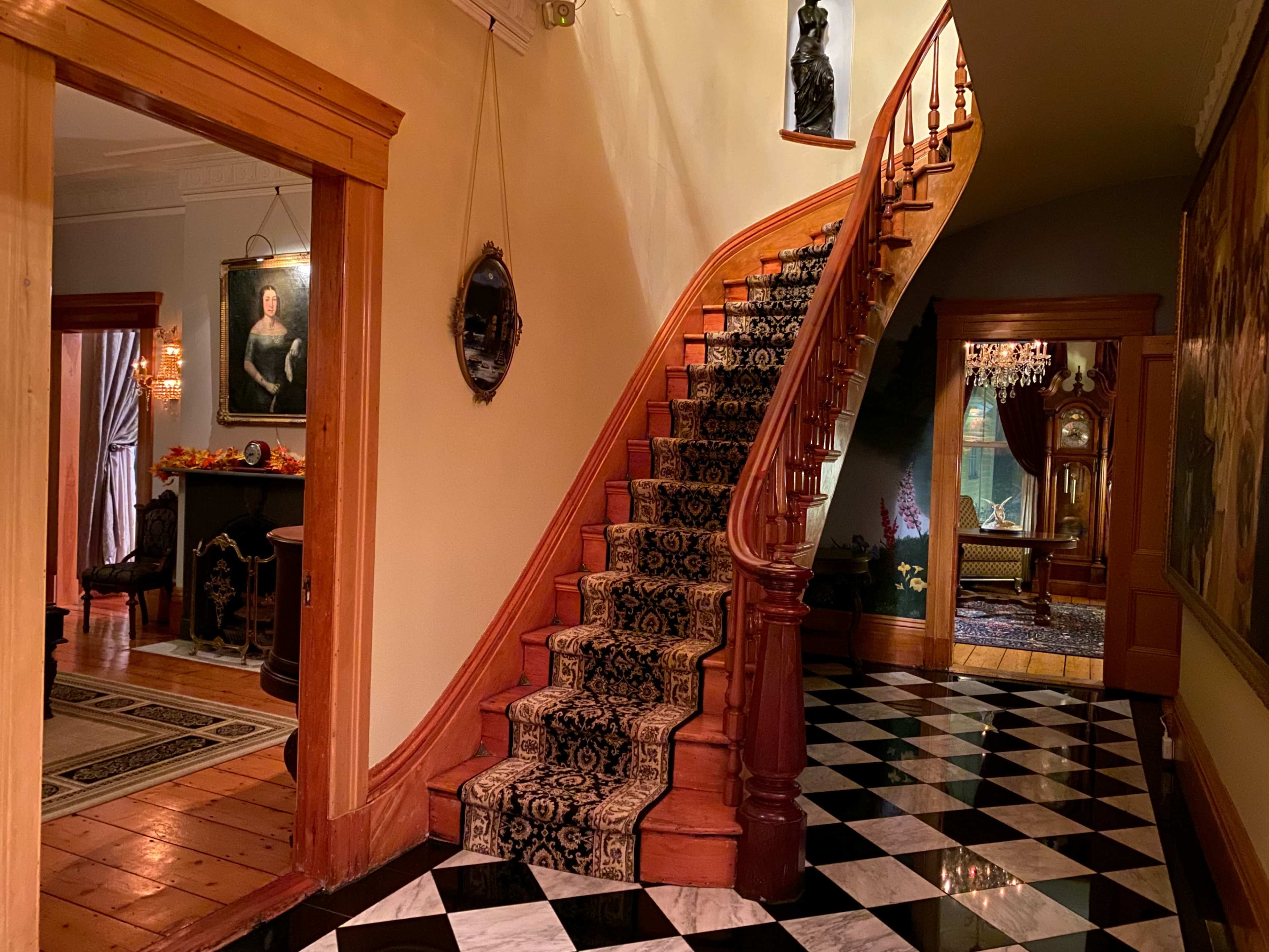 The image shows a grand foyer with a spiral staircase, adorned with a patterned runner, leading to an upper level, flanked by an ornate floor and various pieces of art.