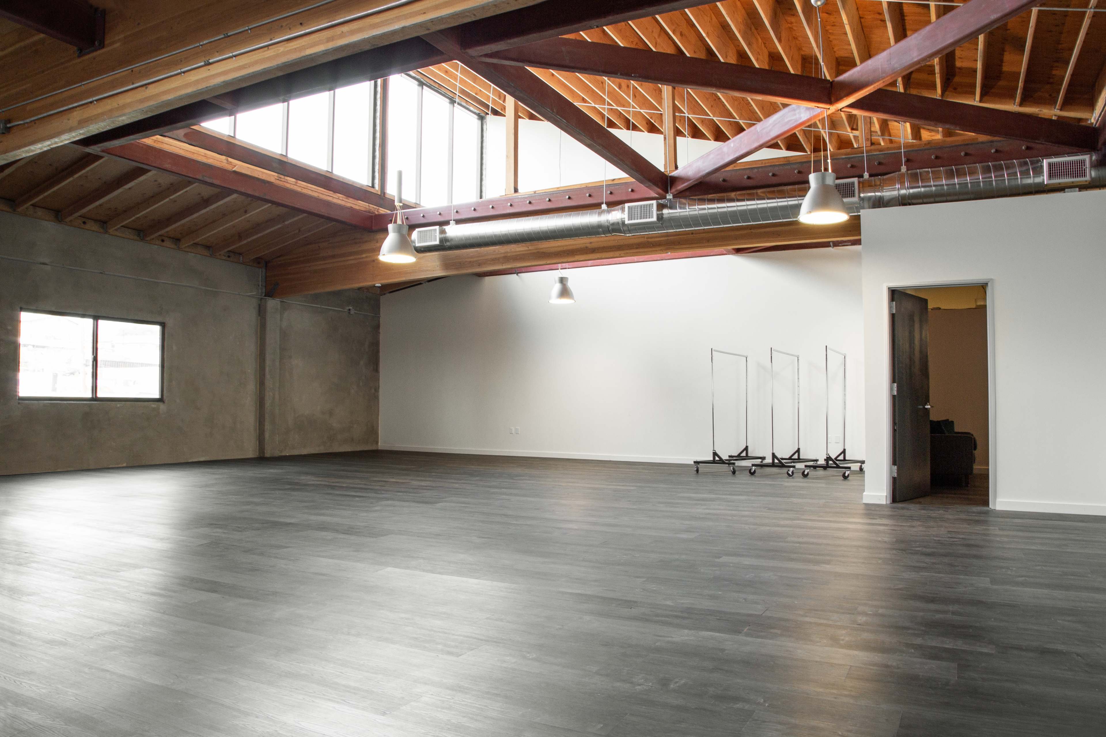 A spacious, empty interior with wooden beams, large windows, and several clothing racks against one wall.
