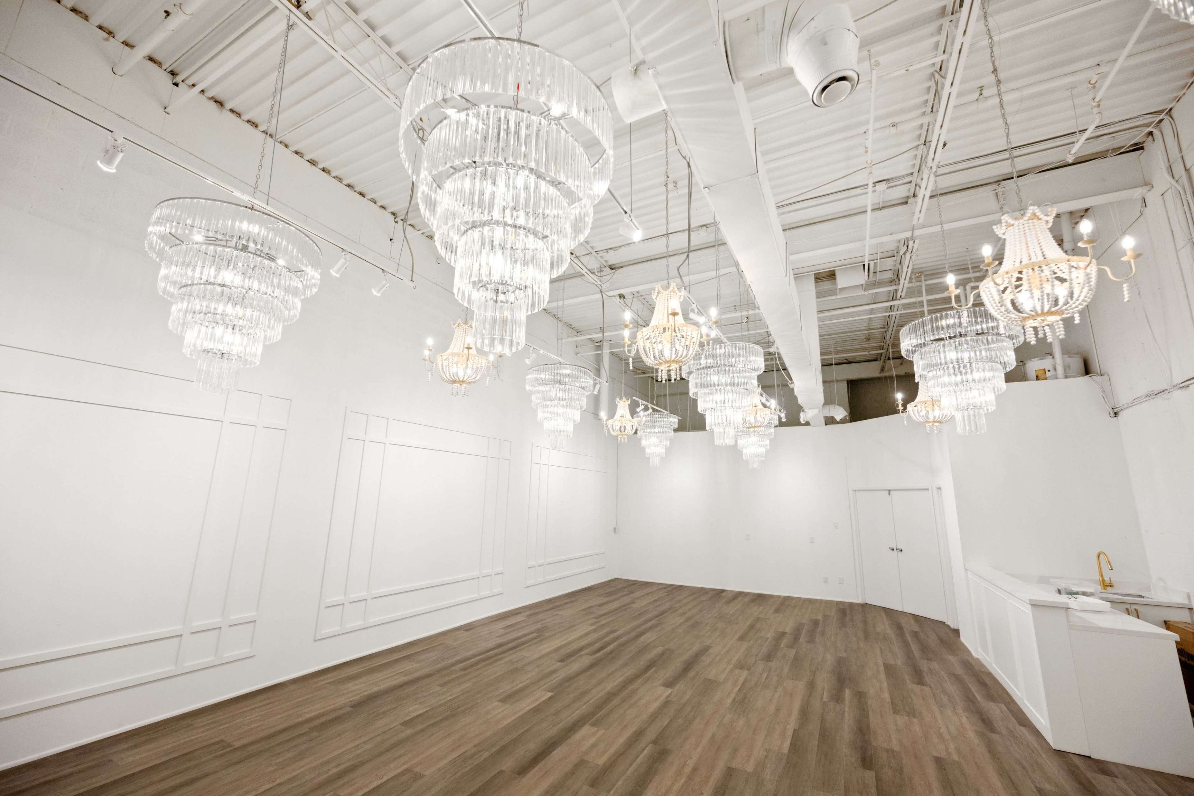 The image shows a spacious, well-lit venue with multiple ornate chandeliers hanging from the ceiling and a wooden floor.