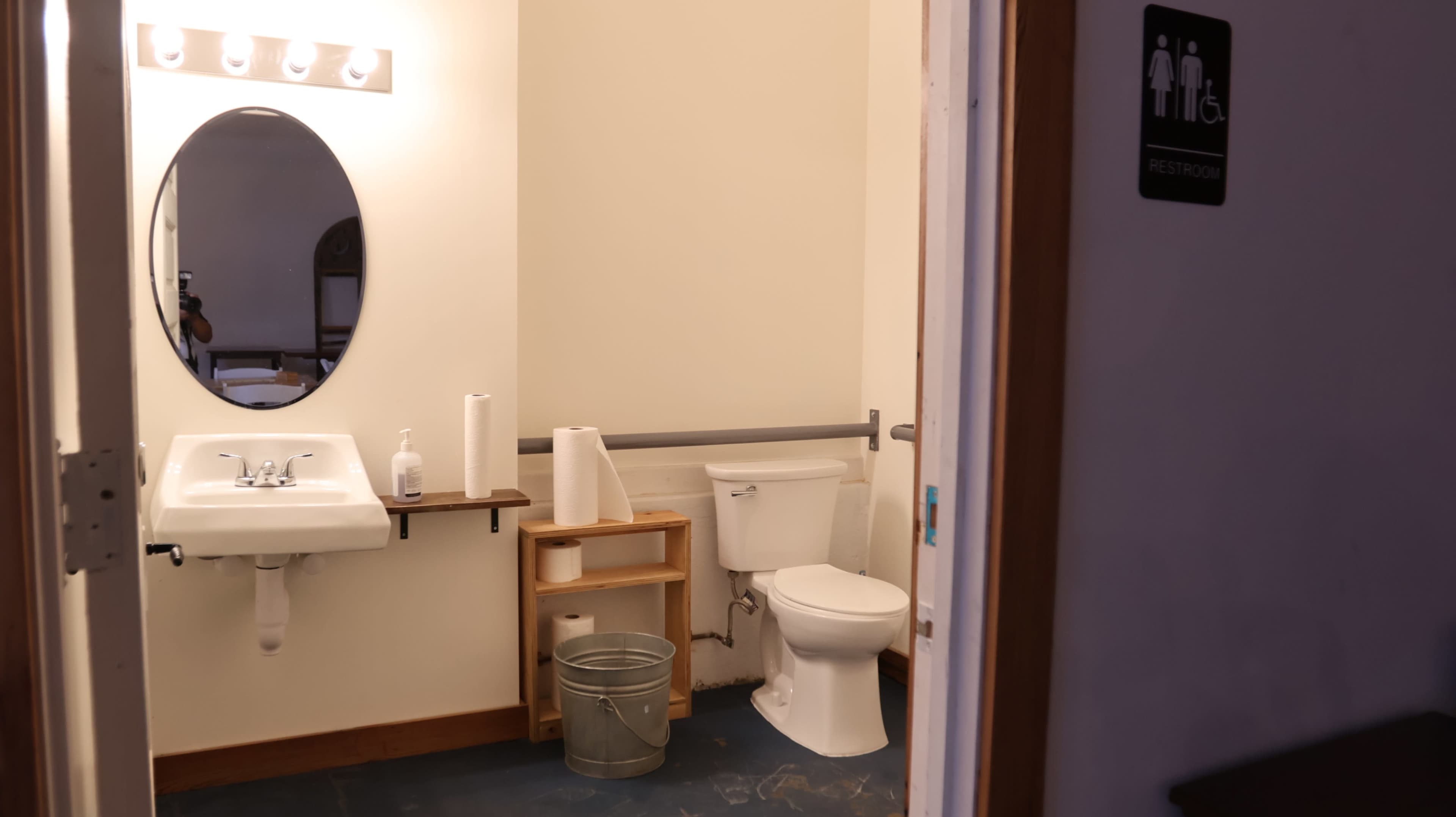The image shows a simple restroom with a sink, toilet, and a small shelf for supplies.