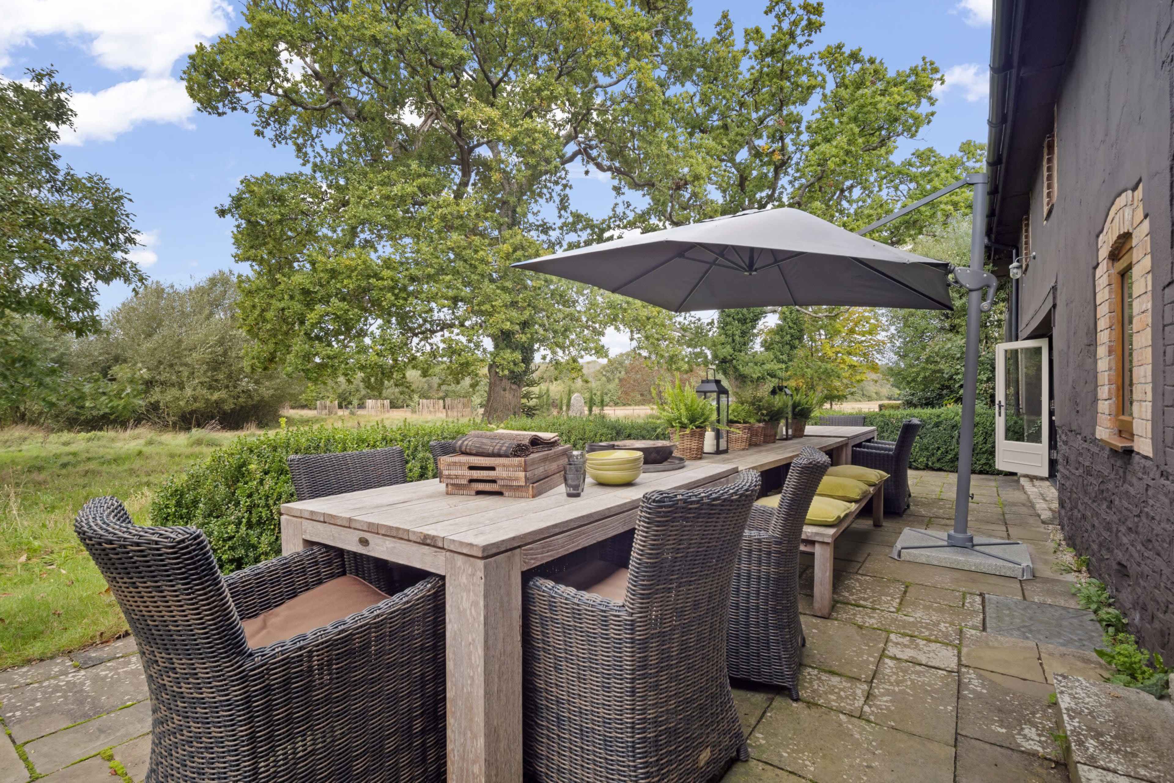 A wooden dining table surrounded by wicker chairs is set on a stone patio under a large umbrella, with trees and greenery in the background.