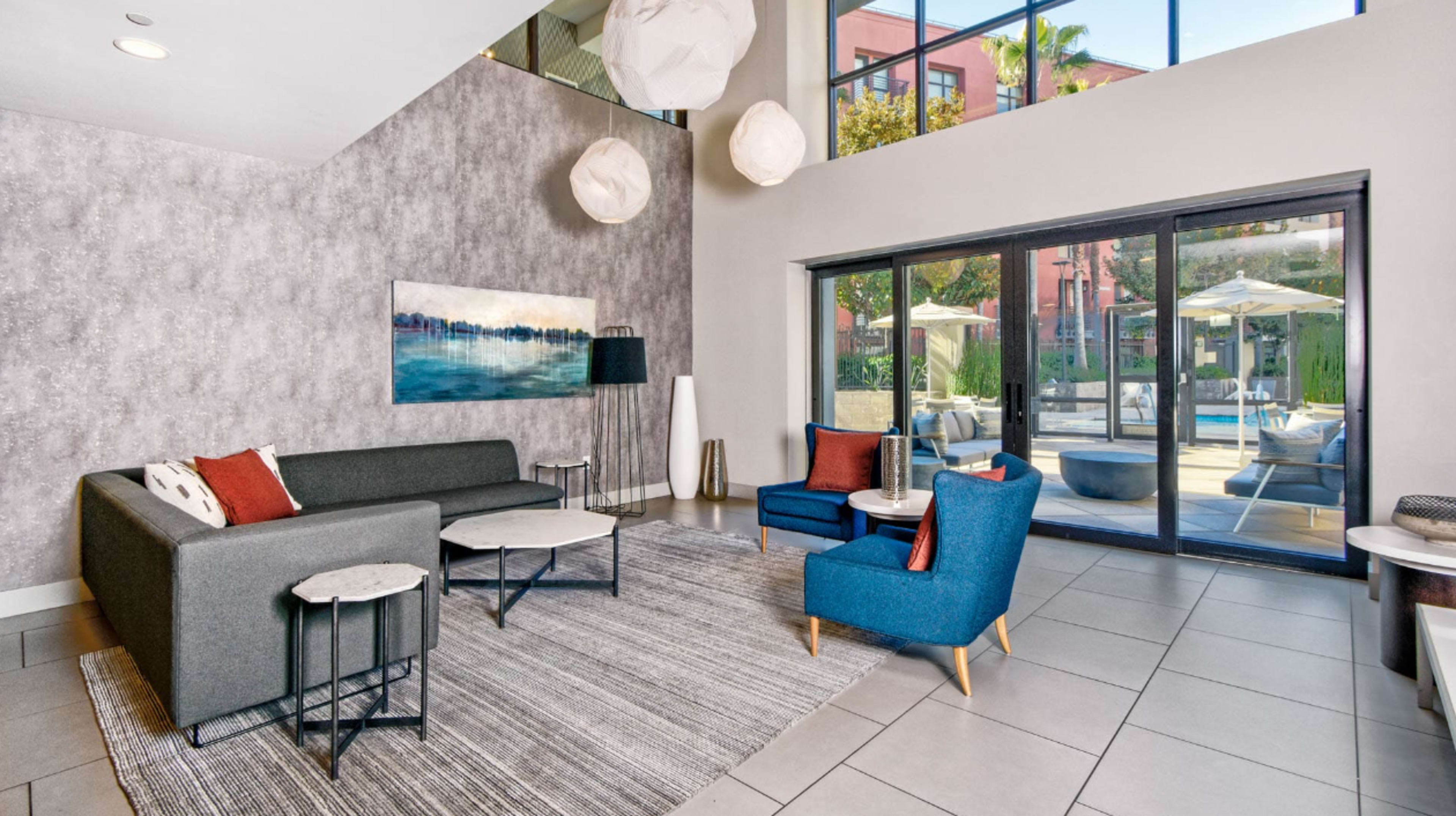 A modern lounge area features gray and blue sofas, round tables, and large windows overlooking a sunny outdoor space with patio umbrellas.