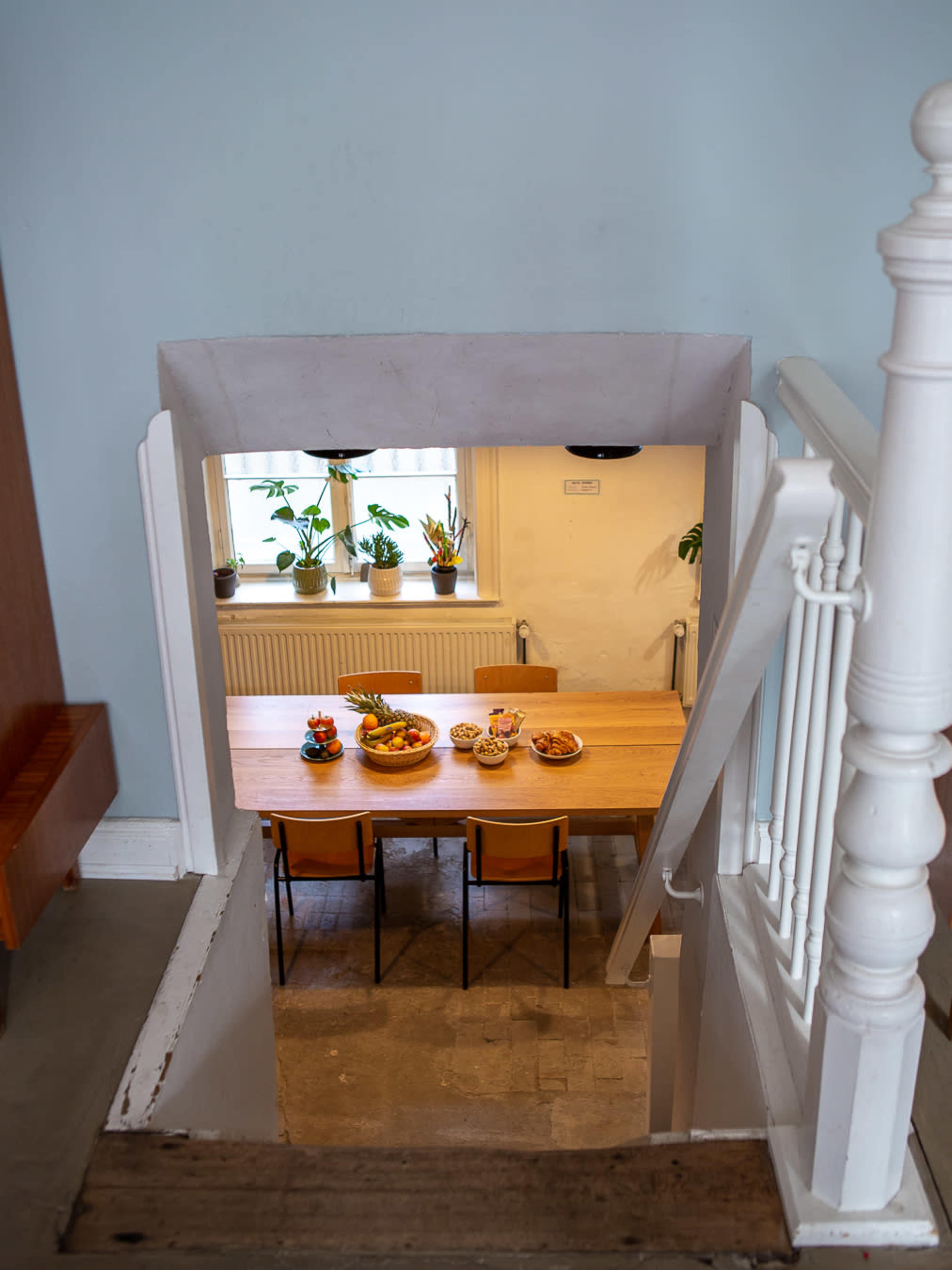 A wooden dining table set with bowls of fruit and surrounded by chairs is visible through an archway at the bottom of a staircase.