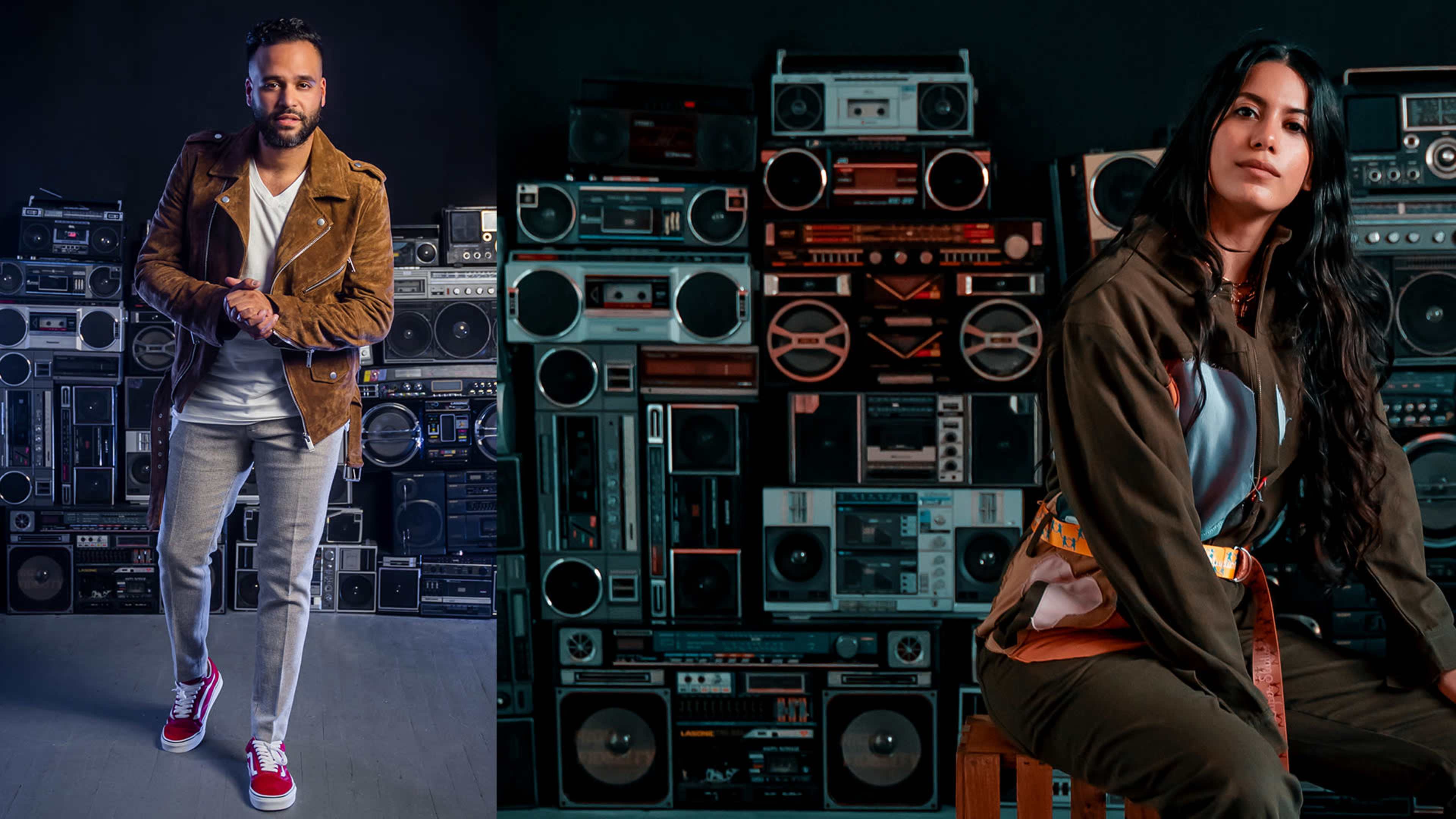 A man in a brown jacket and a woman in a colorful outfit pose in front of a wall adorned with vintage boomboxes.