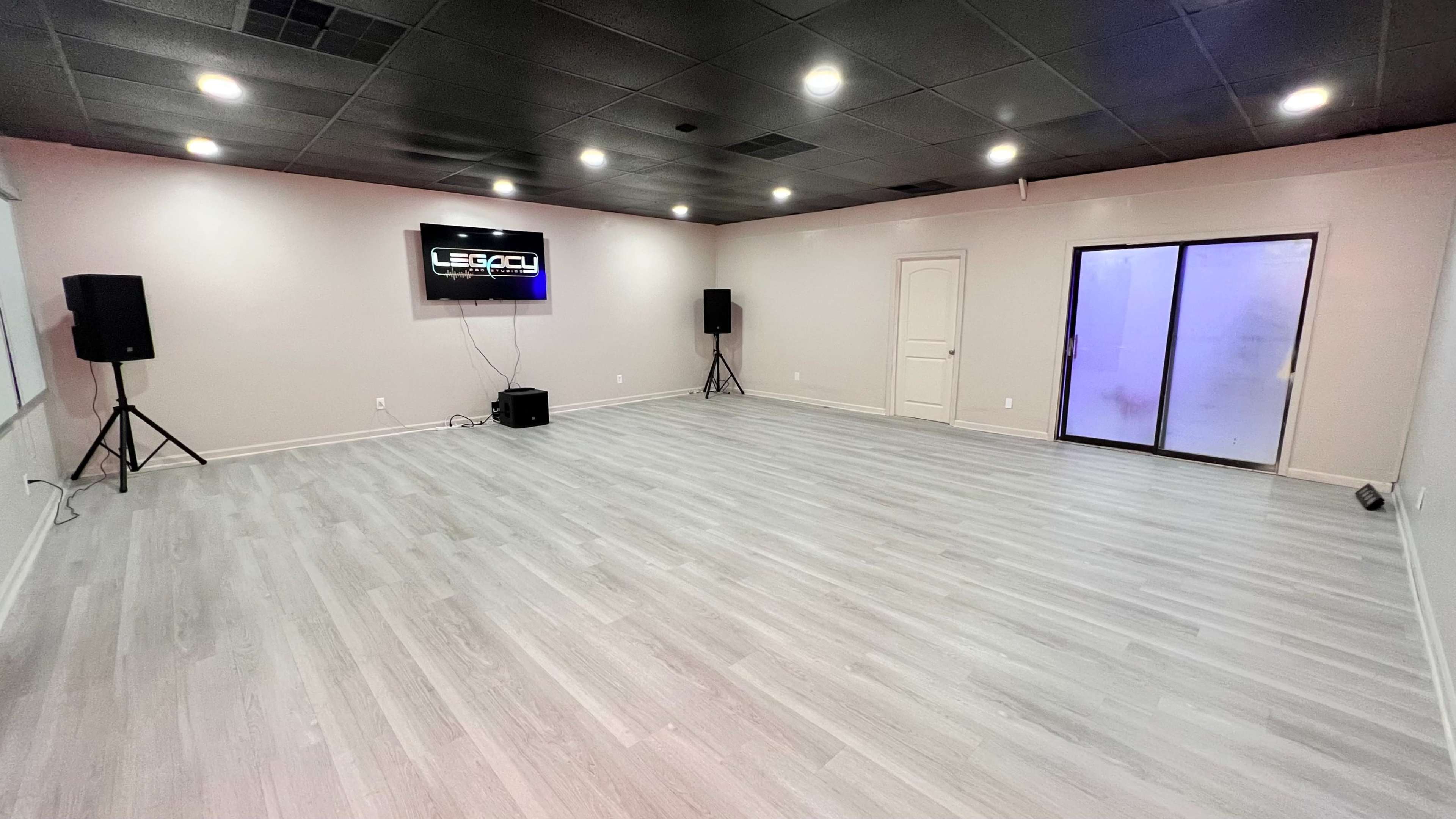 The image shows an empty room with gray wooden flooring, a wall-mounted screen displaying the word "LEGACY," two floor speakers, and a set of double doors leading outside.
