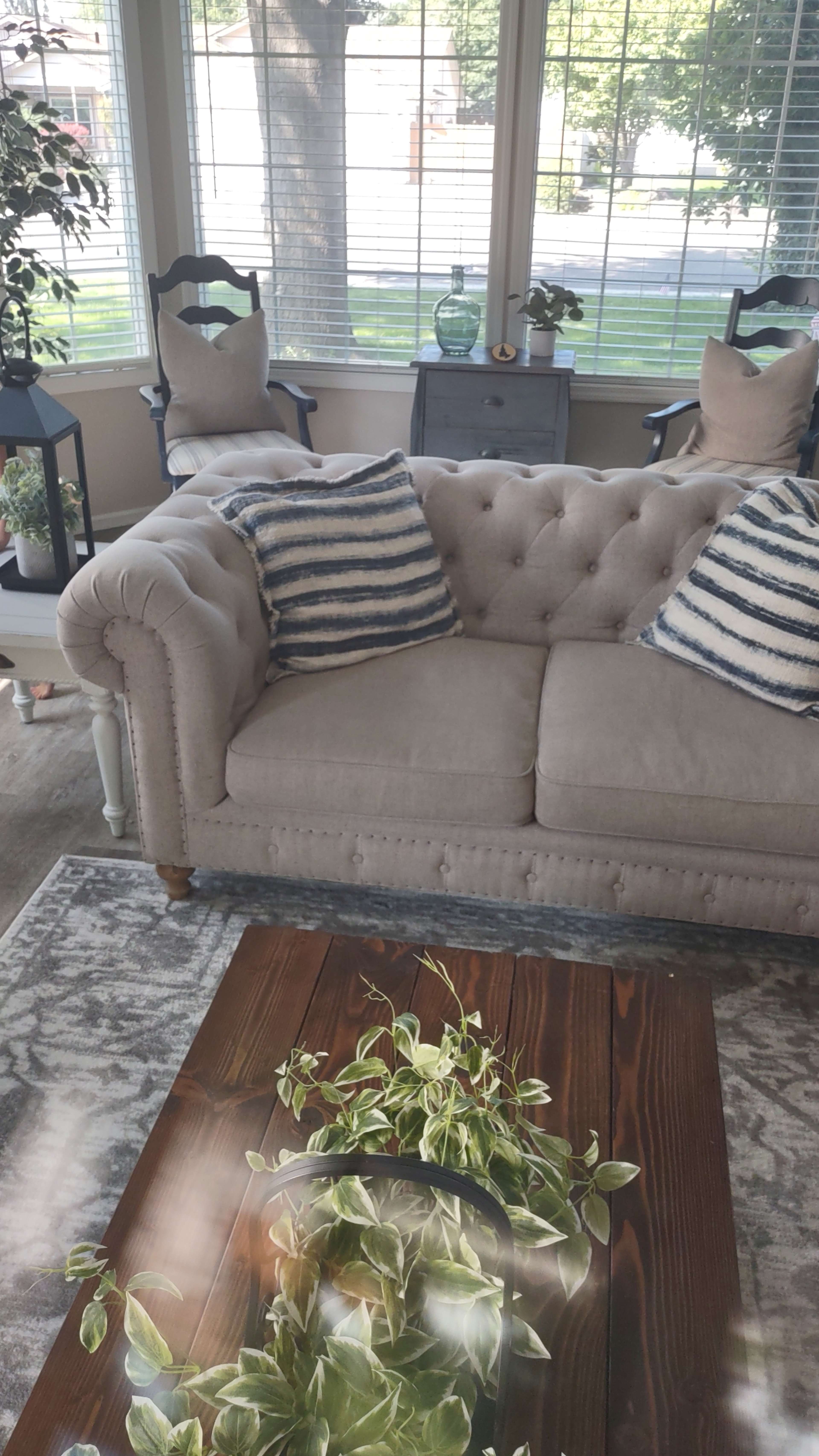 A light-colored tufted couch with striped pillows, positioned in a sunlit room with large windows, accompanied by a wooden coffee table adorned with greenery.