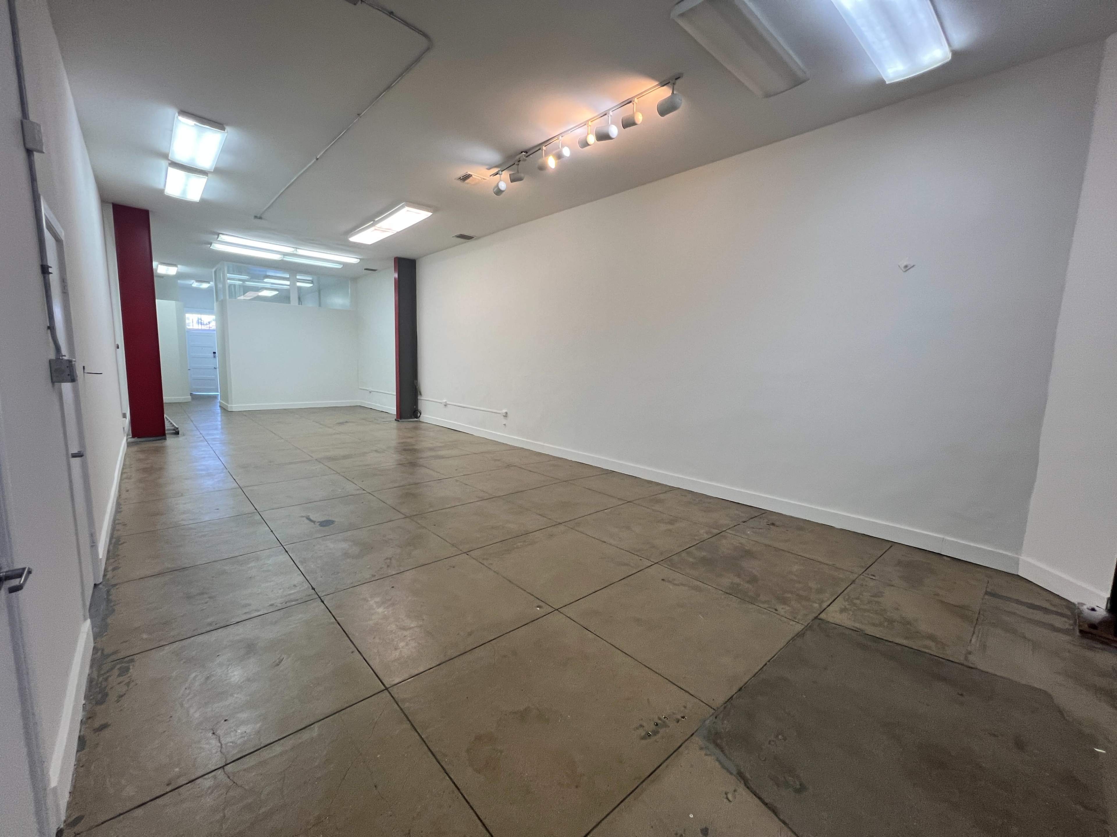 The image shows a spacious, empty room with concrete floors and white walls, featuring ceiling lights and a red accent wall at one end.