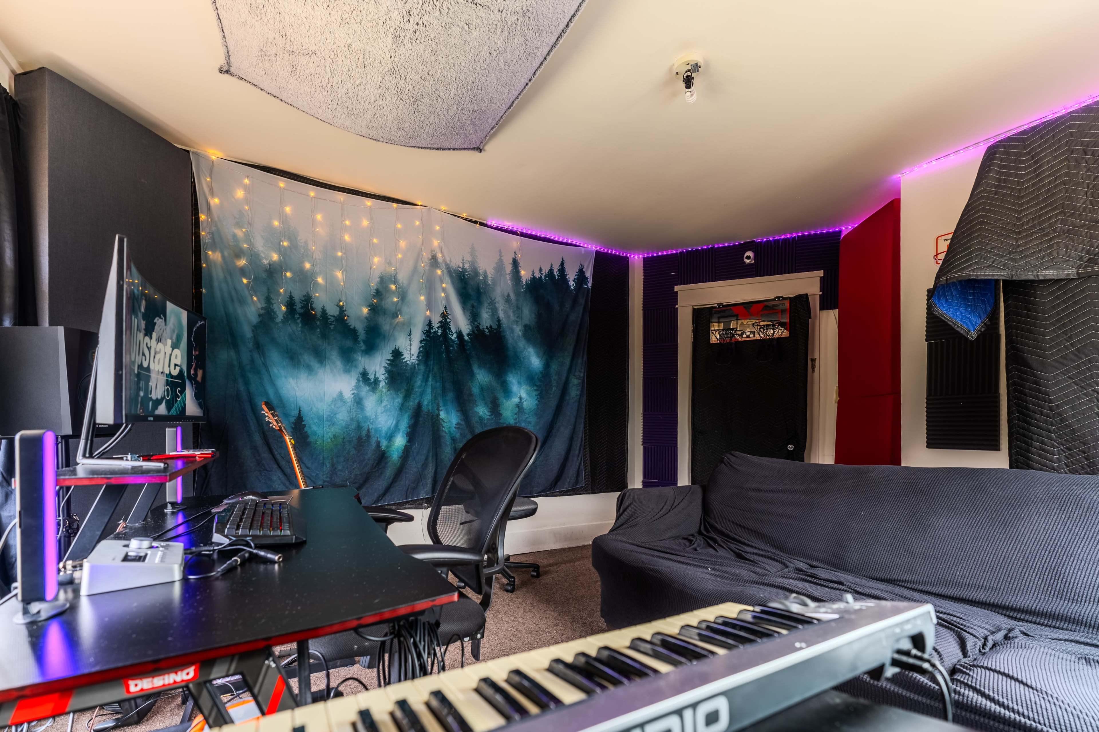 A home studio features a keyboard, a desk with two monitors, and a wall adorned with a forest backdrop and string lights.