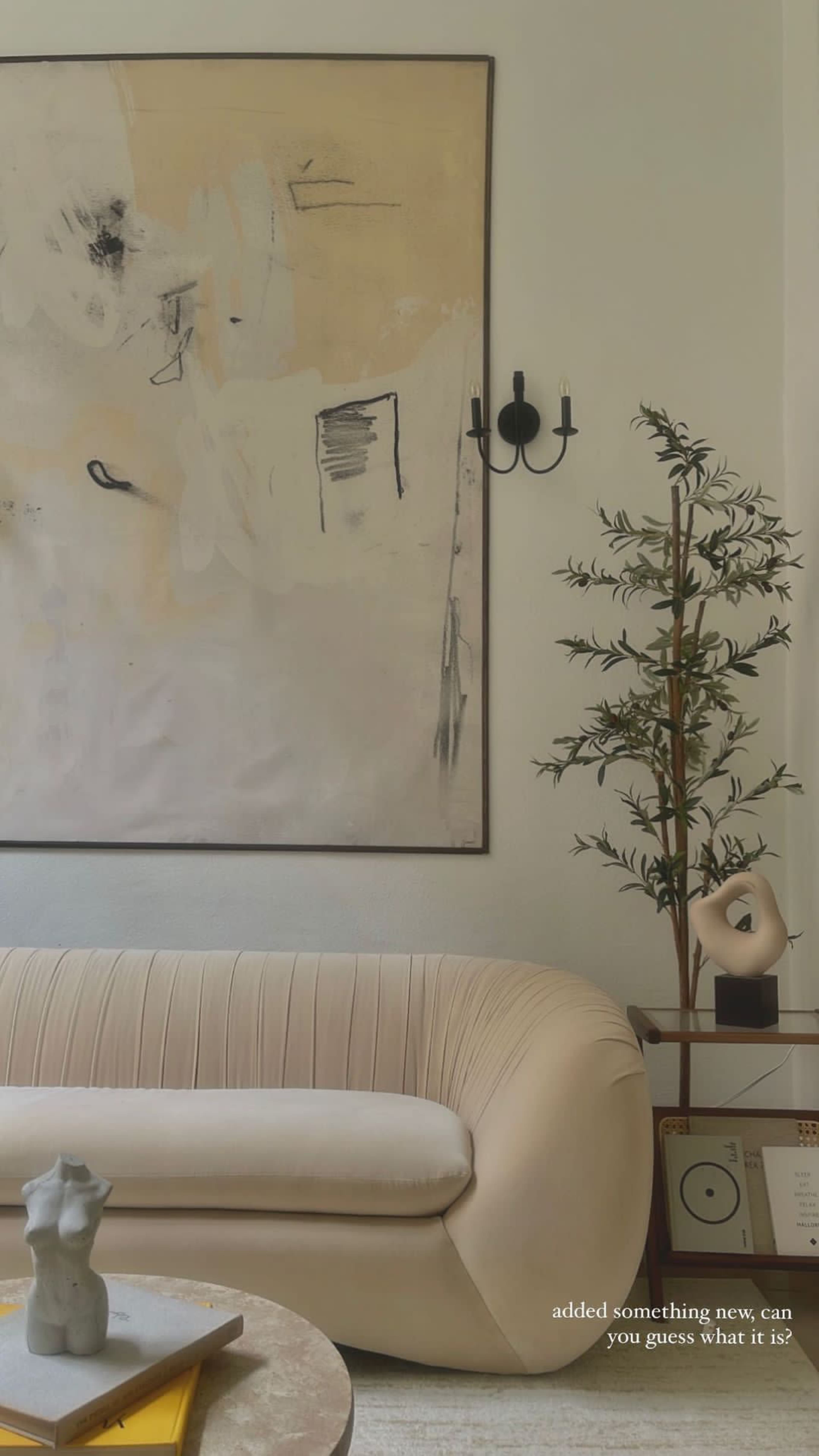 A light-colored, curved sofa sits in a room with a large abstract painting on the wall and a decorative plant in the corner.