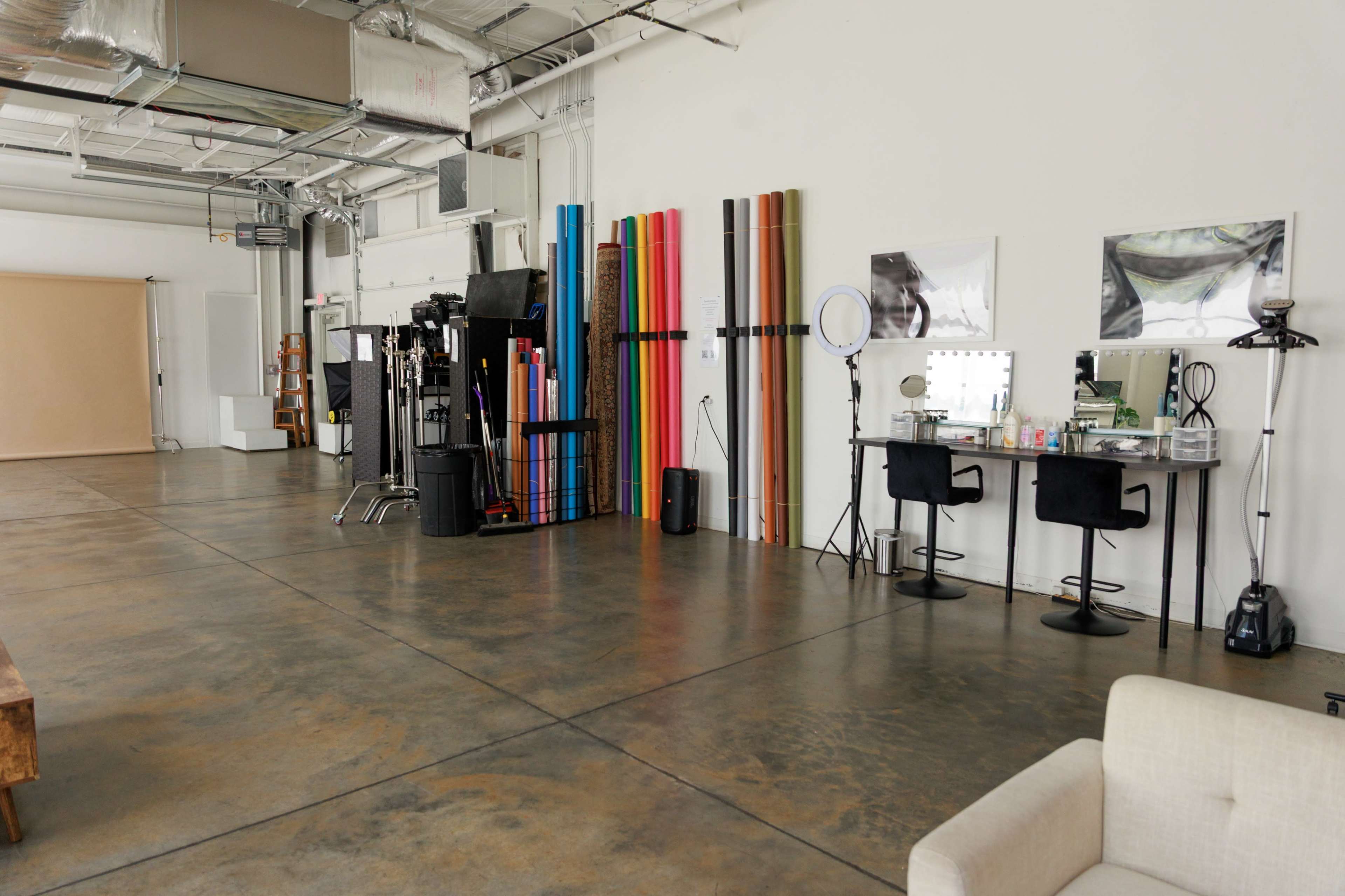The image shows a spacious studio interior with colorful backdrops on one wall, two makeup stations with mirrors, and a sofa in the foreground.