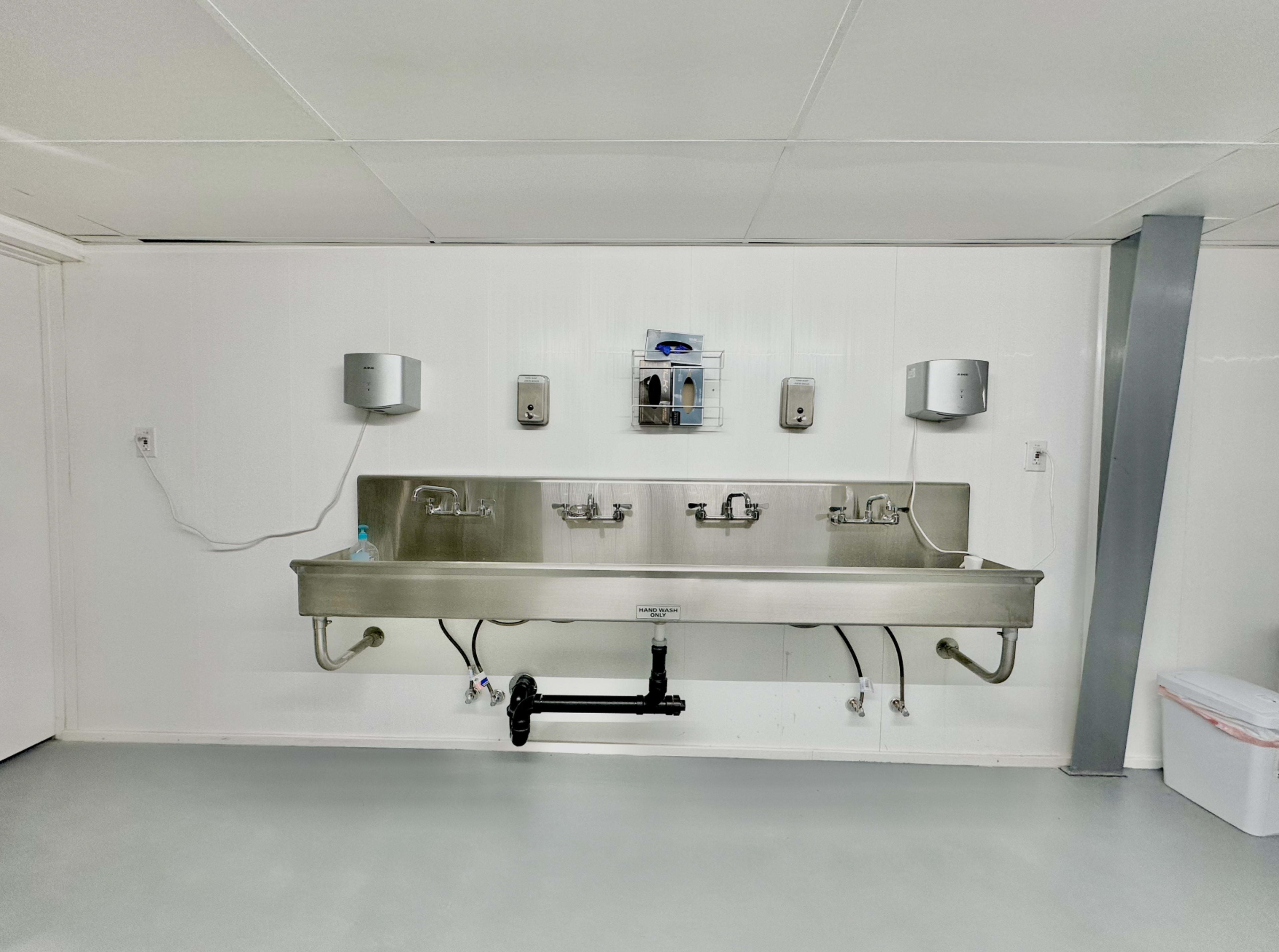 The image shows a stainless steel utility sink mounted on a white wall, equipped with faucets and soap dispensers, in a plain room with a gray floor.